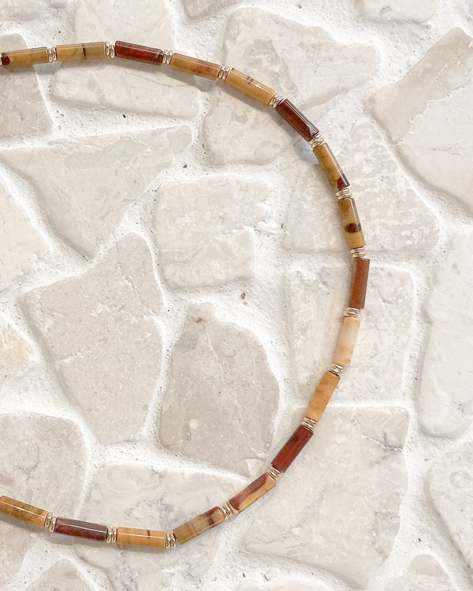 A beaded necklace made of cylindrical stone beads in tan, brown, and reddish tones, arranged in a circle on a light stone tile surface.