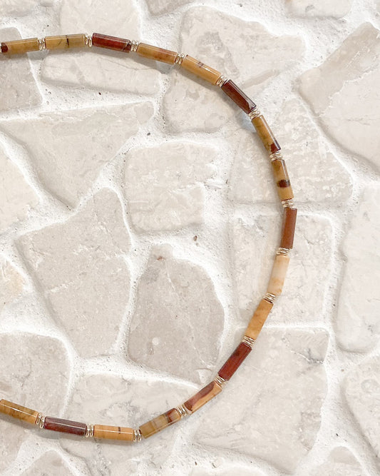 A beaded necklace made of cylindrical stone beads in tan, brown, and reddish tones, arranged in a circle on a light stone tile surface.