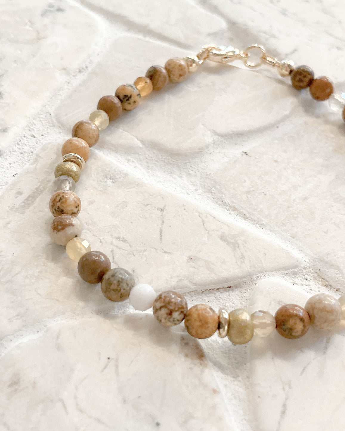 A beaded bracelet made of round natural stone beads in earthy brown, beige, and cream tones, with a small white bead and a gold clasp, displayed on a light stone tile surface.