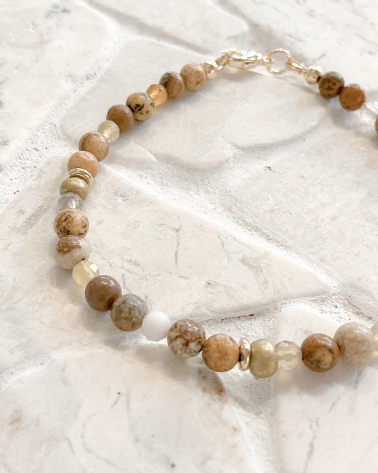 A beaded bracelet made of round natural stone beads in earthy brown, beige, and cream tones, with a small white bead and a gold clasp, displayed on a light stone tile surface.