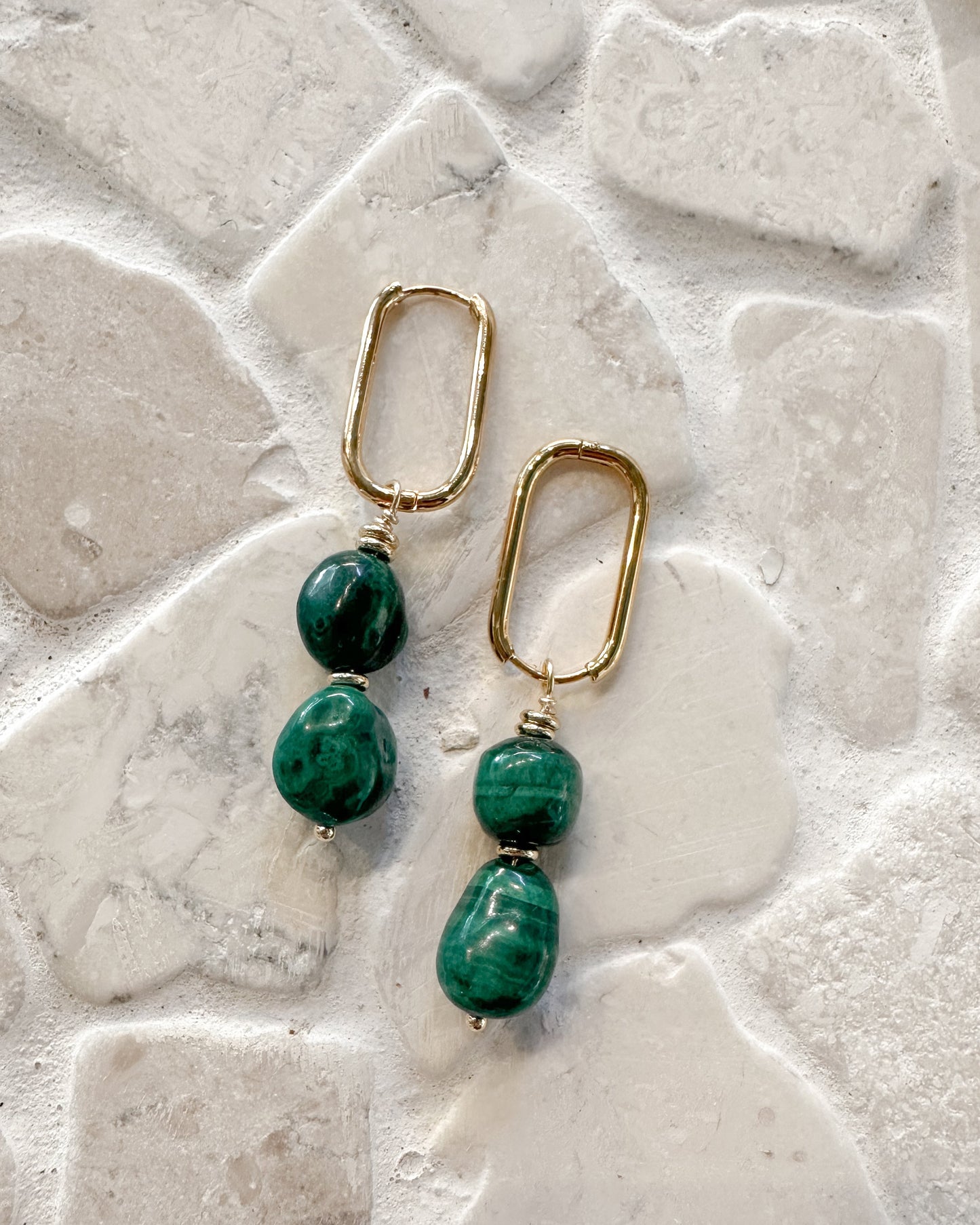 A pair of gold drop earrings with green malachite beads, displayed on a textured light gray stone tile background.