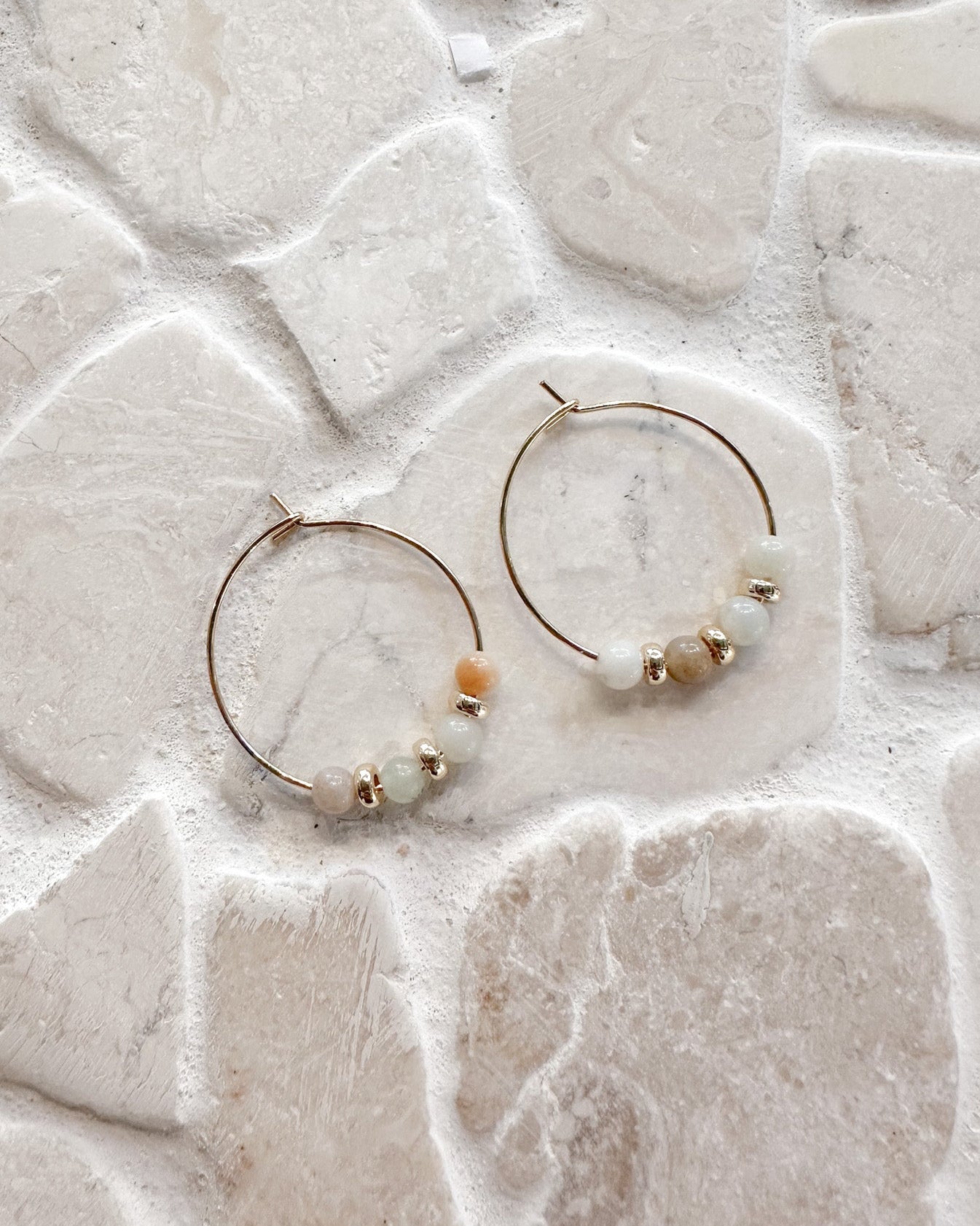 A pair of gold hoop earrings with small beads, displayed on a textured light beige stone tile surface.