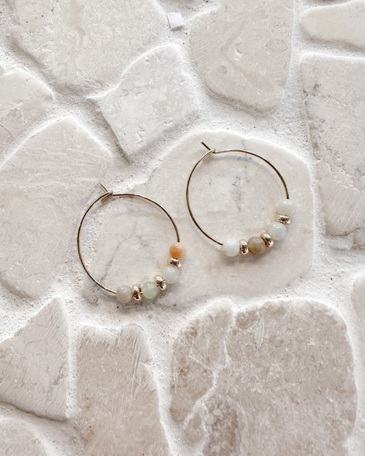 A pair of gold hoop earrings with small beads, displayed on a textured light beige stone tile surface.