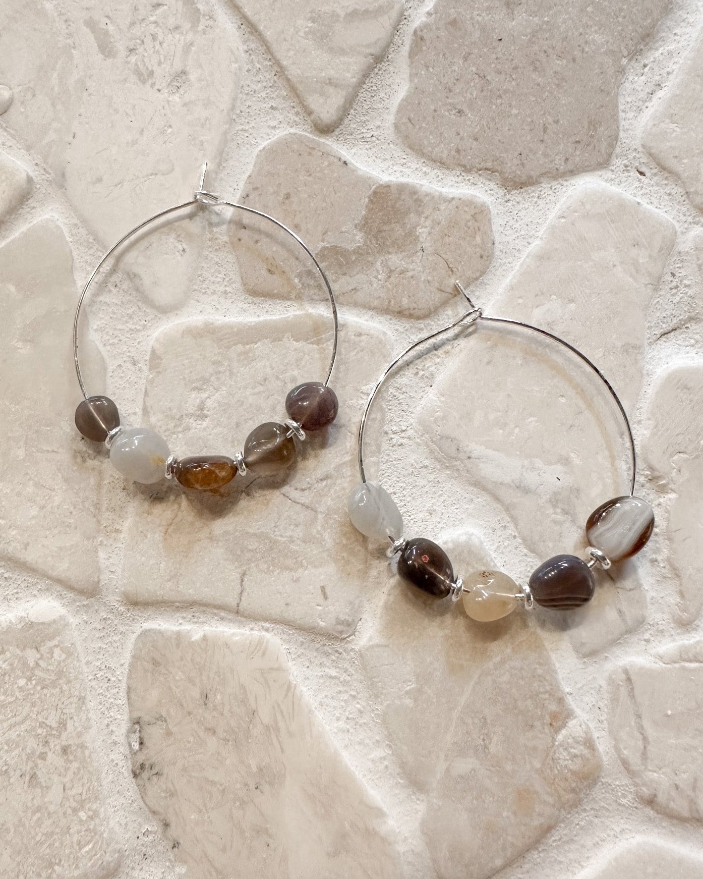 A pair of silver hoop earrings with small faceted beads in brown, cream, and gray, displayed on a light beige stone tile background.