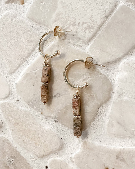A pair of handmade earrings with gold-toned hoops and textured brown beads, displayed on a light beige stone surface.