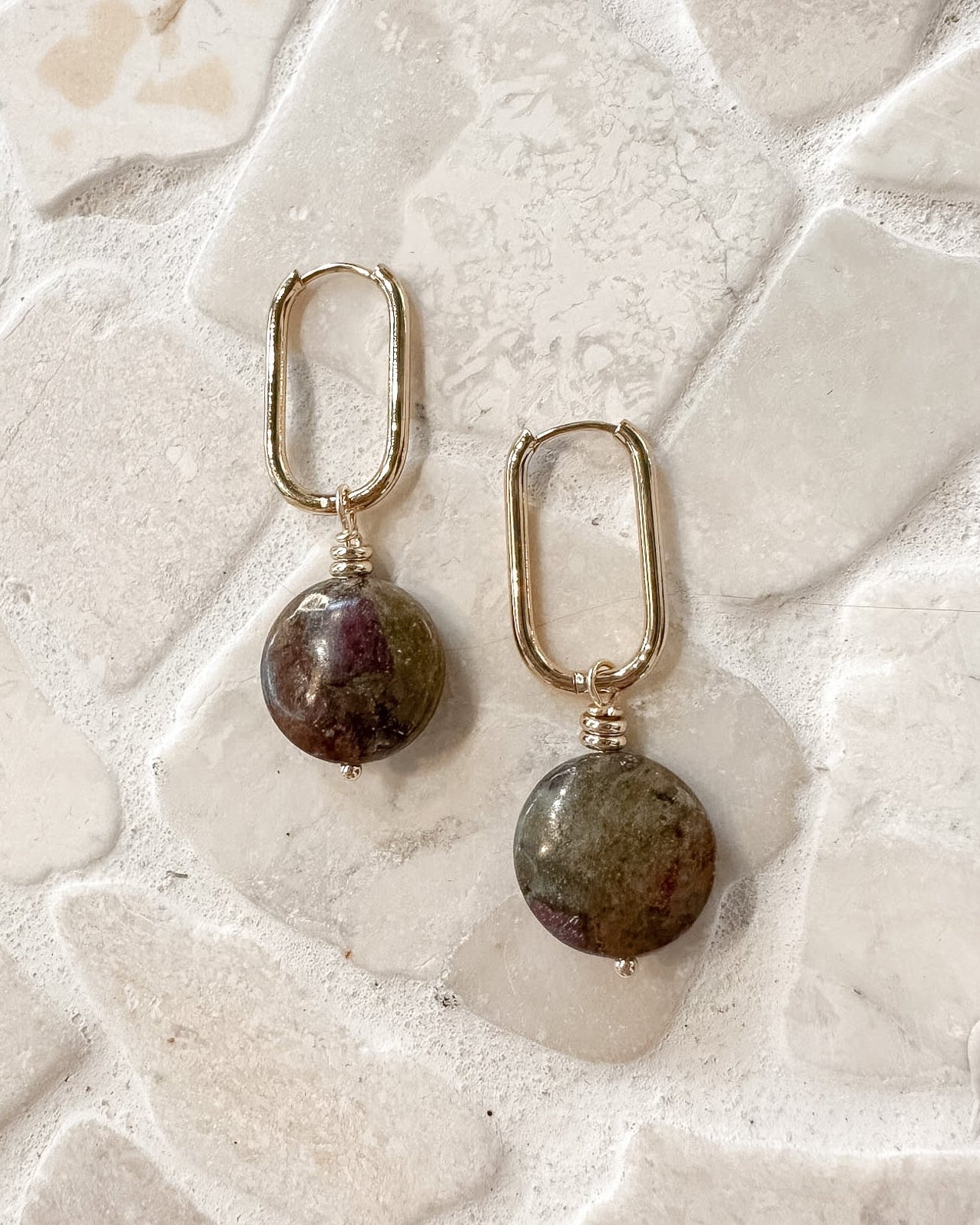 A pair of gold-toned drop earrings with oval-shaped natural stone pendants, displayed on a textured, light-colored stone surface.