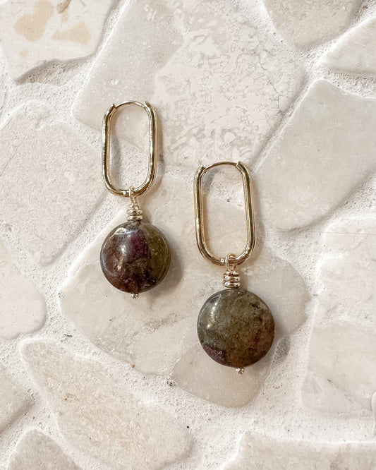 A pair of gold-toned drop earrings with oval-shaped natural stone pendants, displayed on a textured, light-colored stone surface.