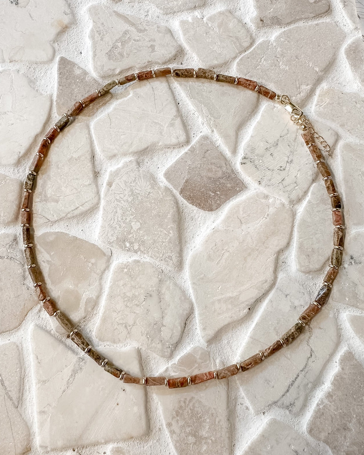 A beaded necklace made of small brown and amber-toned rectangular beads, arranged in a circle on a light beige stone-tile surface.