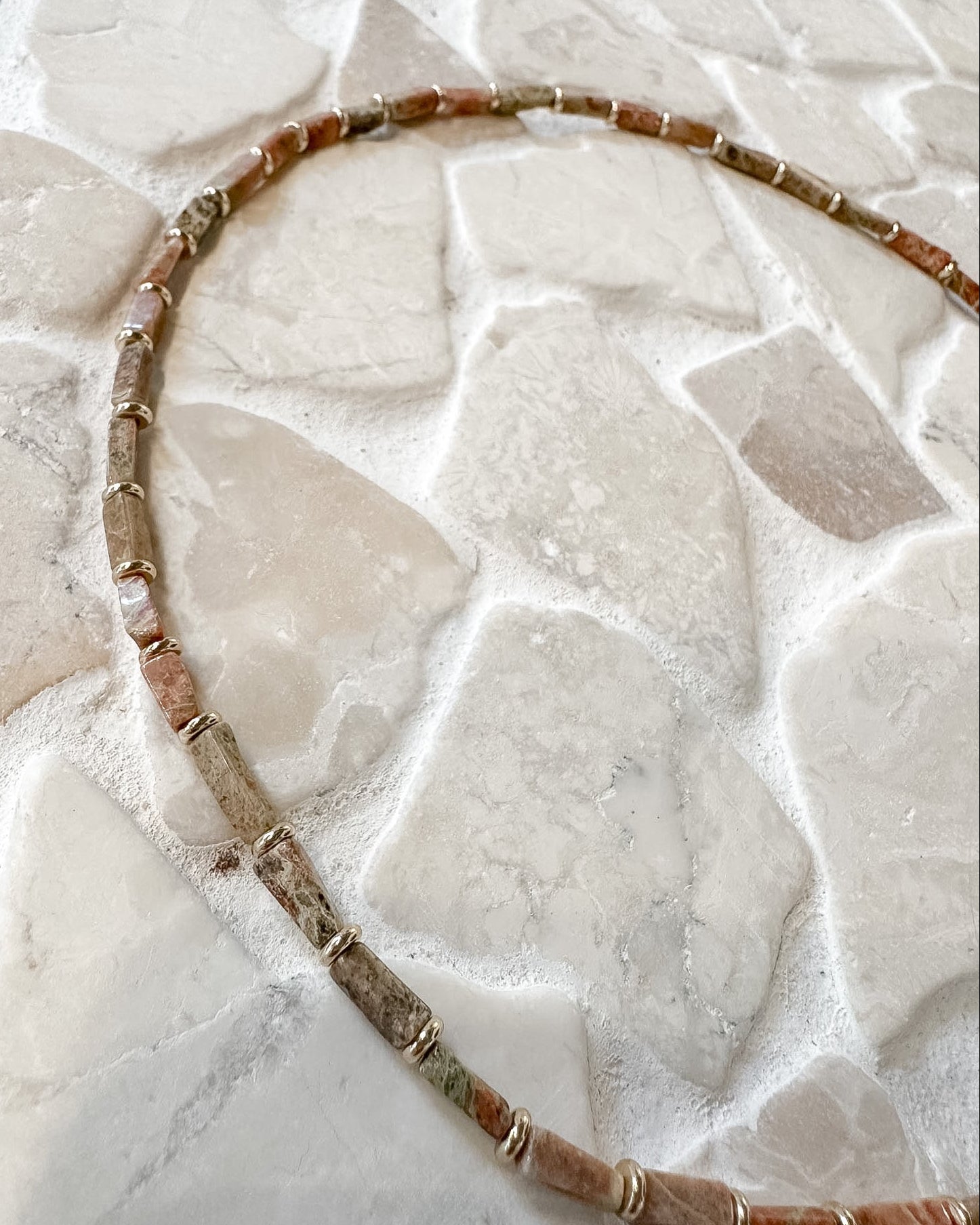 A long beaded necklace made of terracotta-colored stone beads with small metallic spacer rings, laid out in a curved shape on a light stone tile surface.