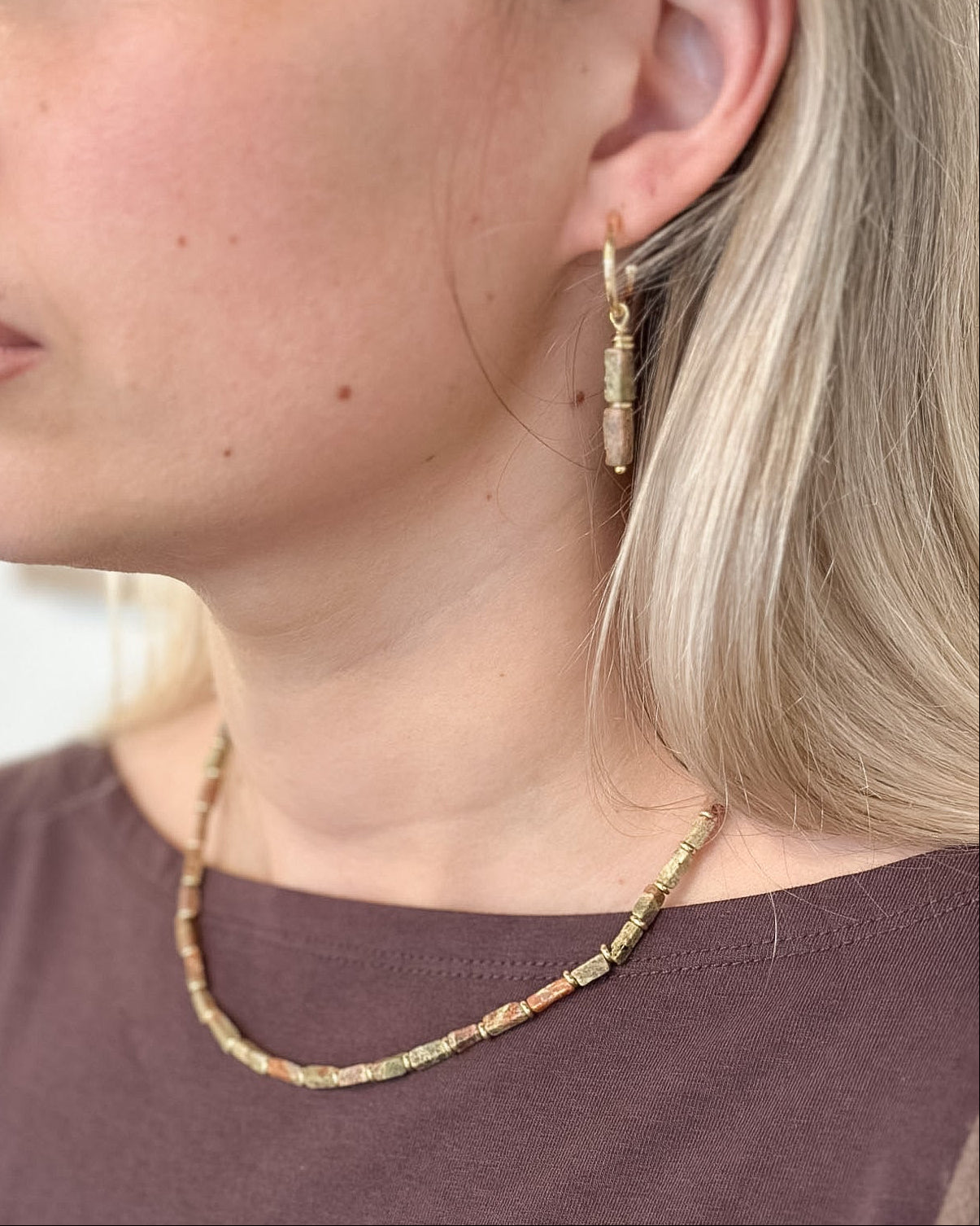 A close-up side profile of a woman wearing a delicate beaded necklace and matching drop hoop earrings. The jewelry features small, multi-colored beads in earthy tones of beige, brown, and muted orange, suspended from gold-toned metal.
