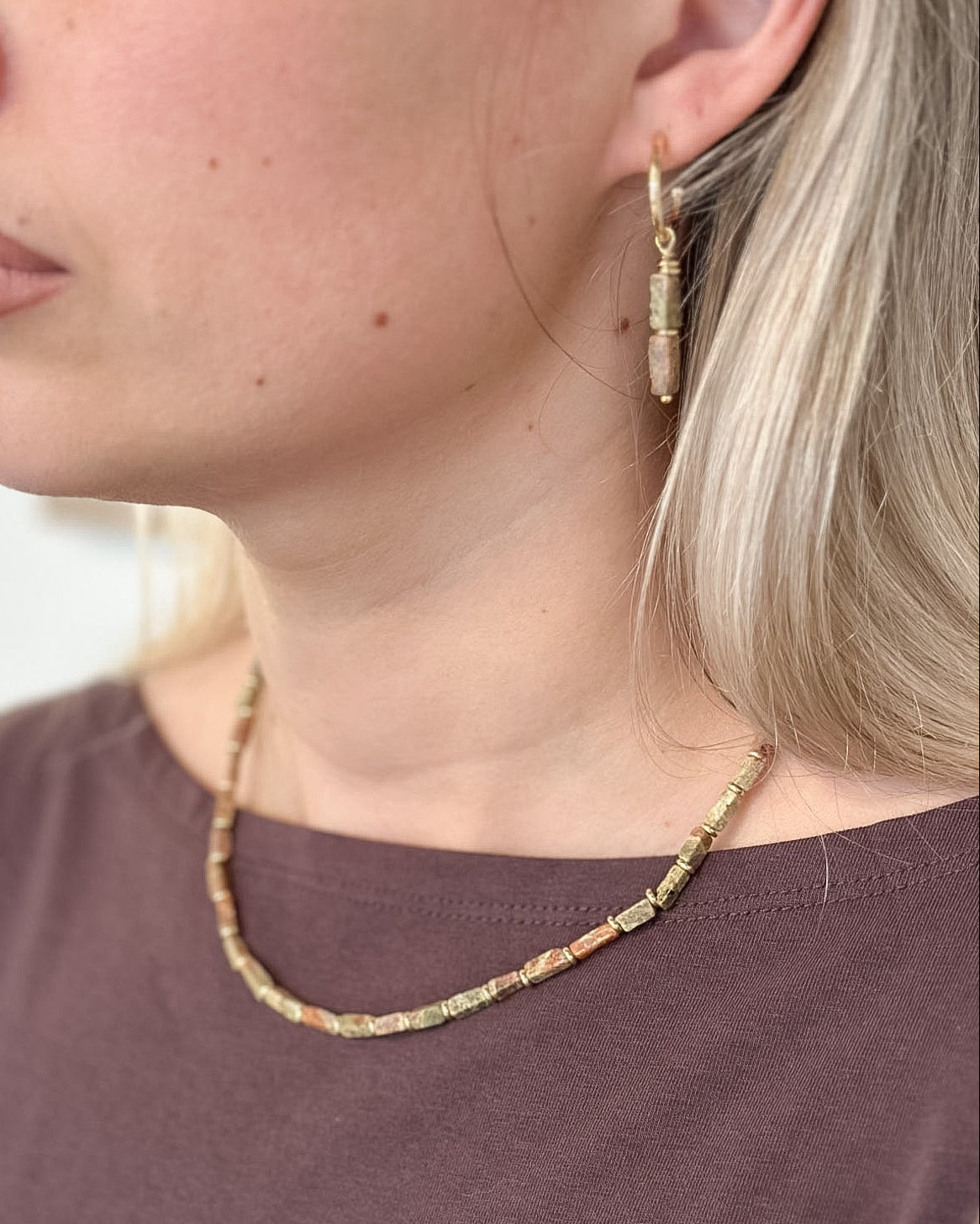 A close-up side profile of a woman wearing a delicate beaded necklace and matching drop hoop earrings. The jewelry features small, multi-colored beads in earthy tones of beige, brown, and muted orange, suspended from gold-toned metal.