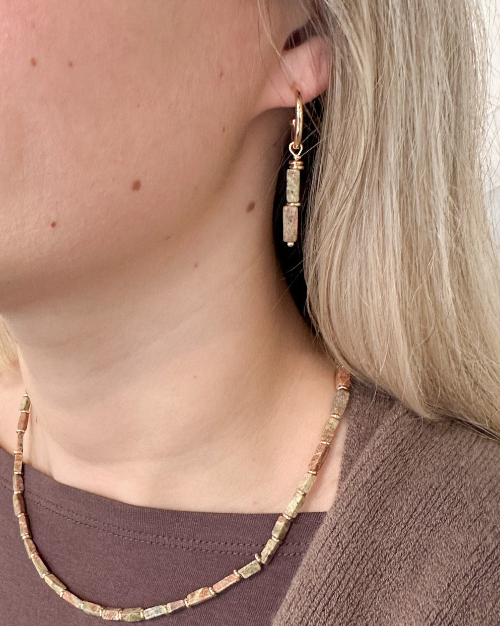A close-up lifestyle shot of a woman wearing a delicate gold-toned beaded necklace and matching drop earrings, paired with a brown knit top.