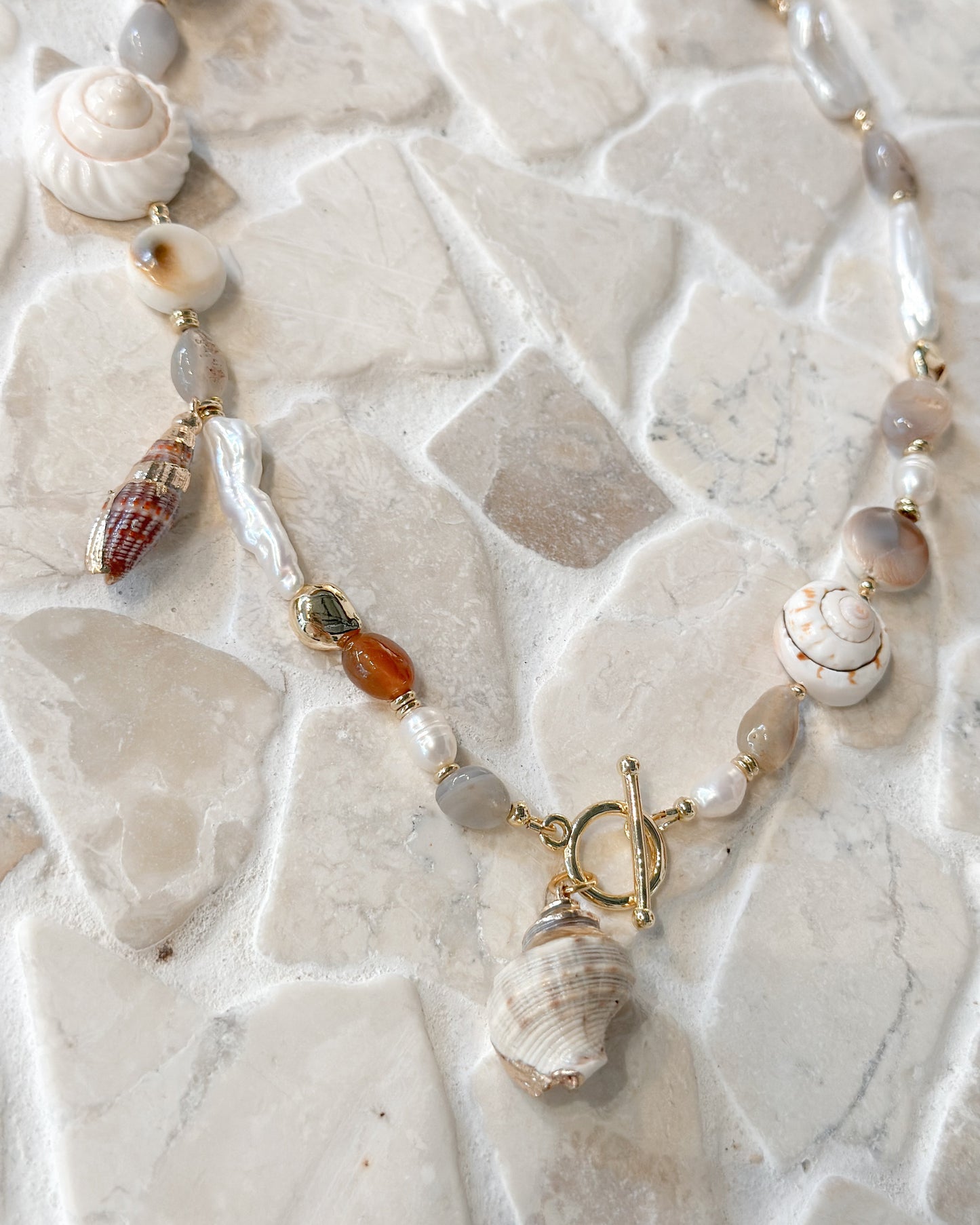 A bohemian-style beaded necklace featuring various seashells, white pearls, and small gold-toned beads, displayed on a light beige stone tile background.