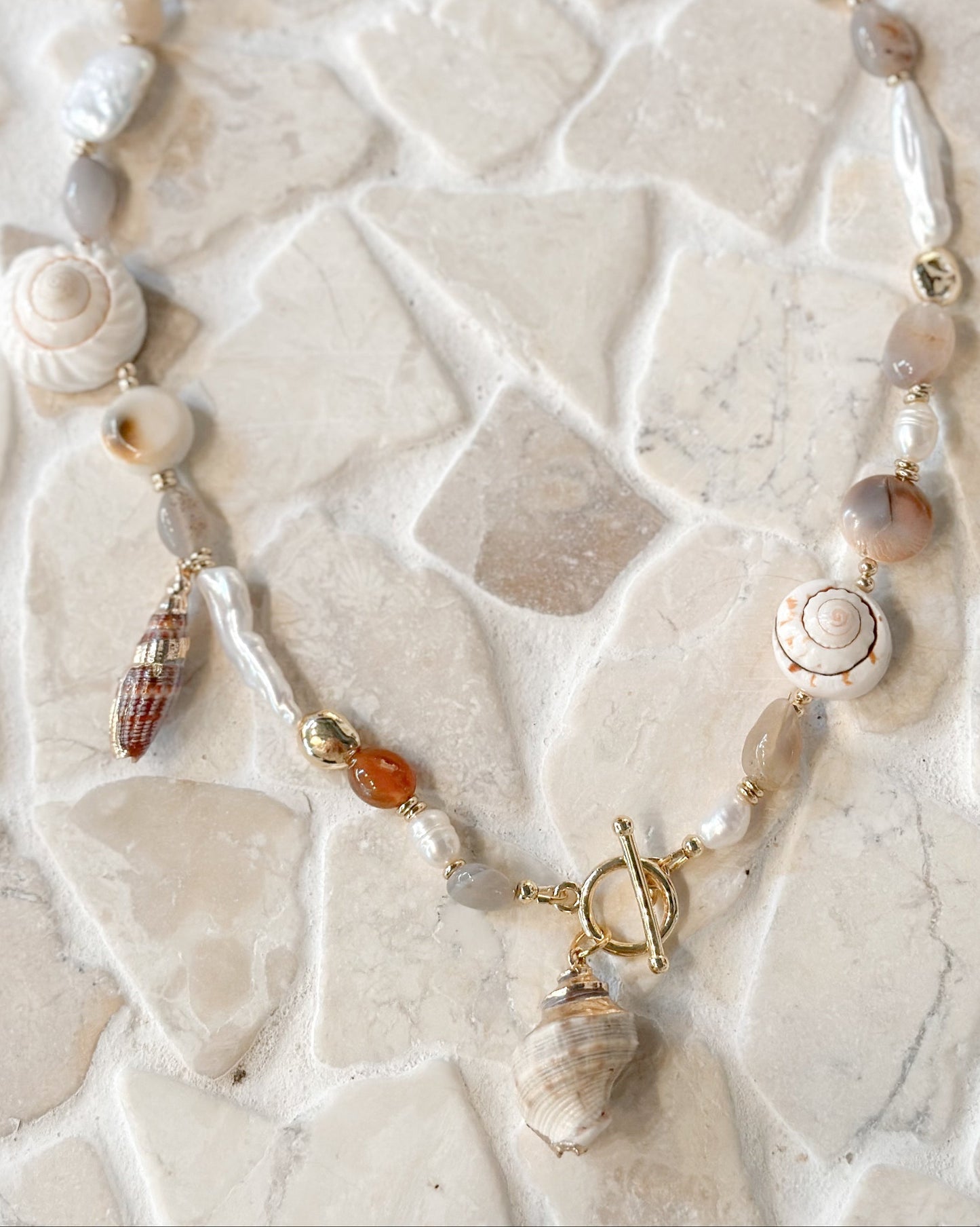 A beaded necklace featuring natural seashells, agate beads in earthy grey and brown tones, and small amber accents, laid flat on a light beige stone tile background.