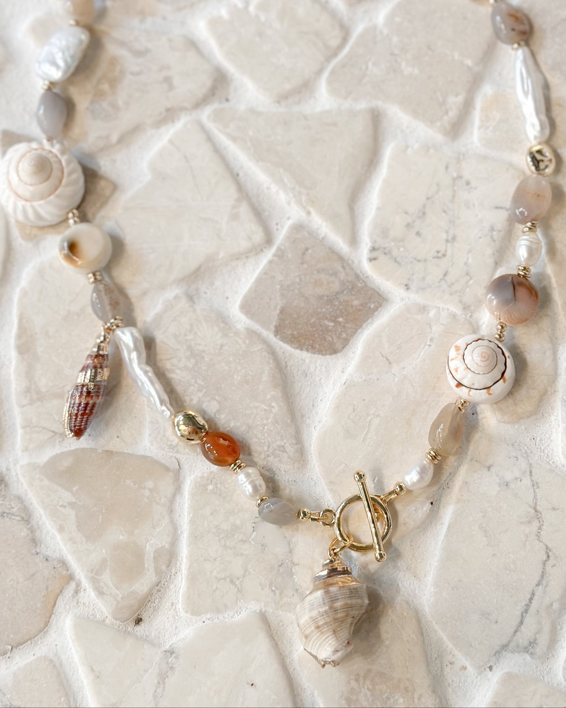 A beaded necklace featuring natural seashells, agate beads in earthy grey and brown tones, and small amber accents, laid flat on a light beige stone tile background.