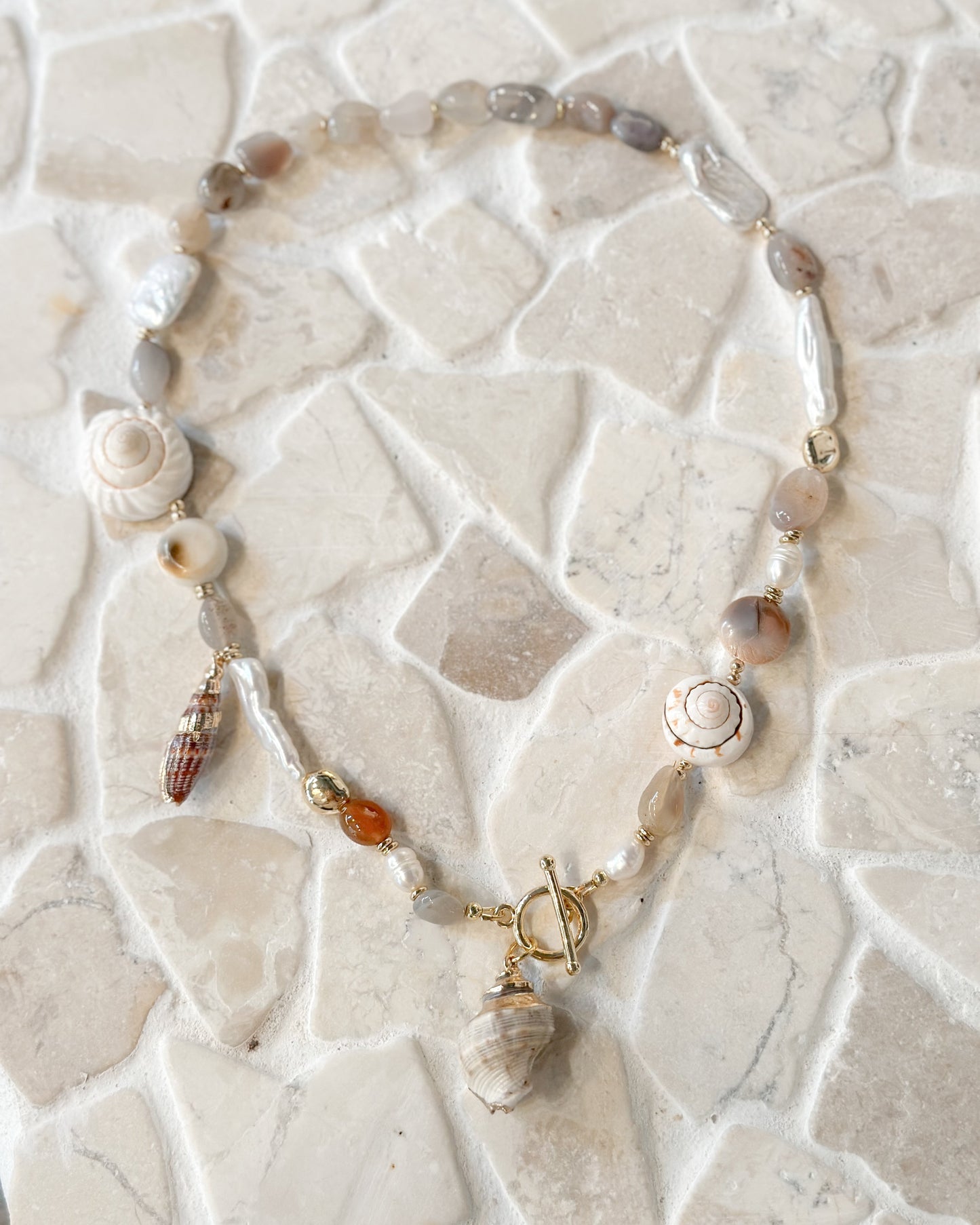 A bohemian-style beaded necklace featuring seashells, baroque pearls, and small amber beads, displayed on a light beige mosaic tile surface.