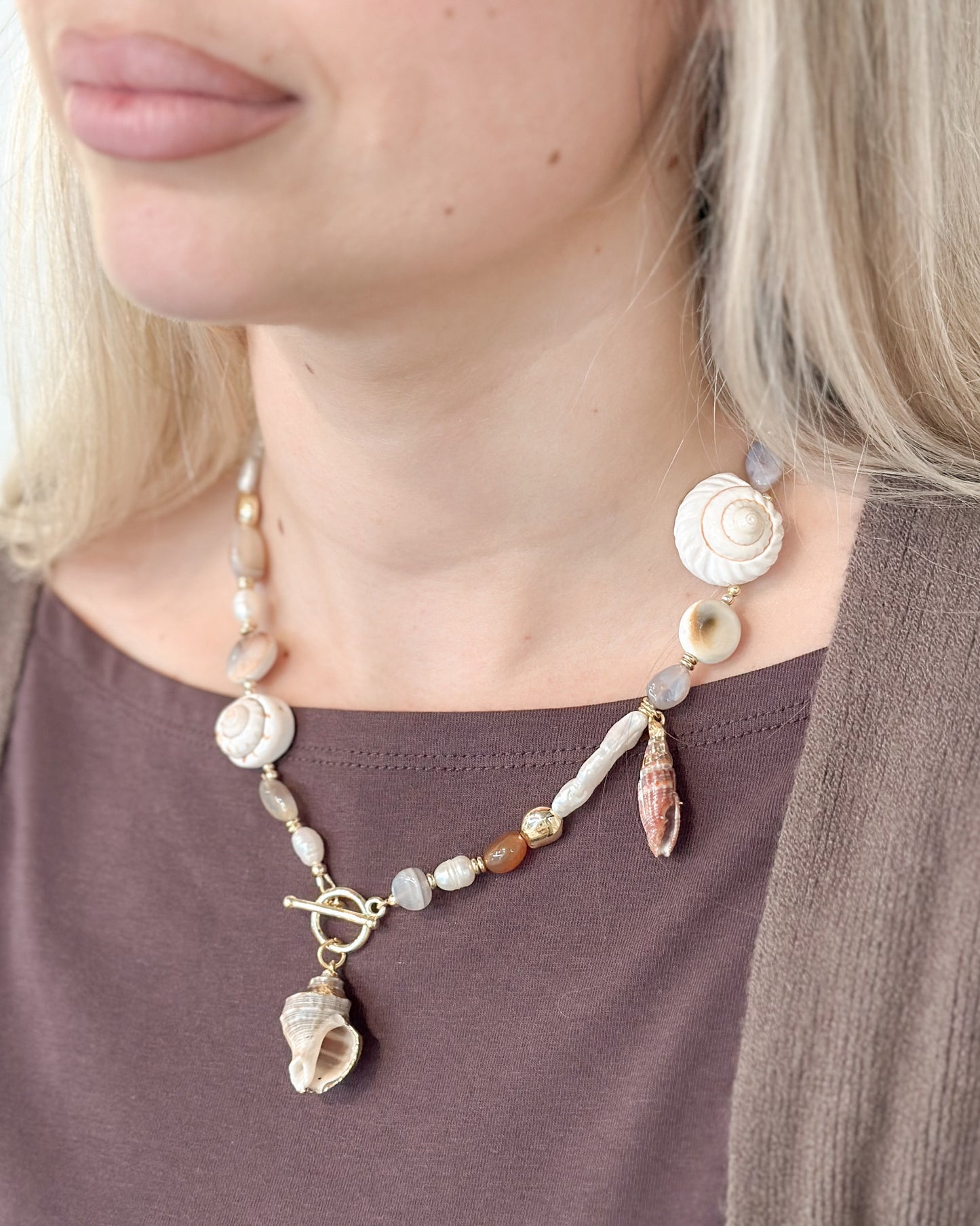 A close-up of a woman wearing a bohemian-style seashell necklace with a toggle clasp over a brown top and cardigan.