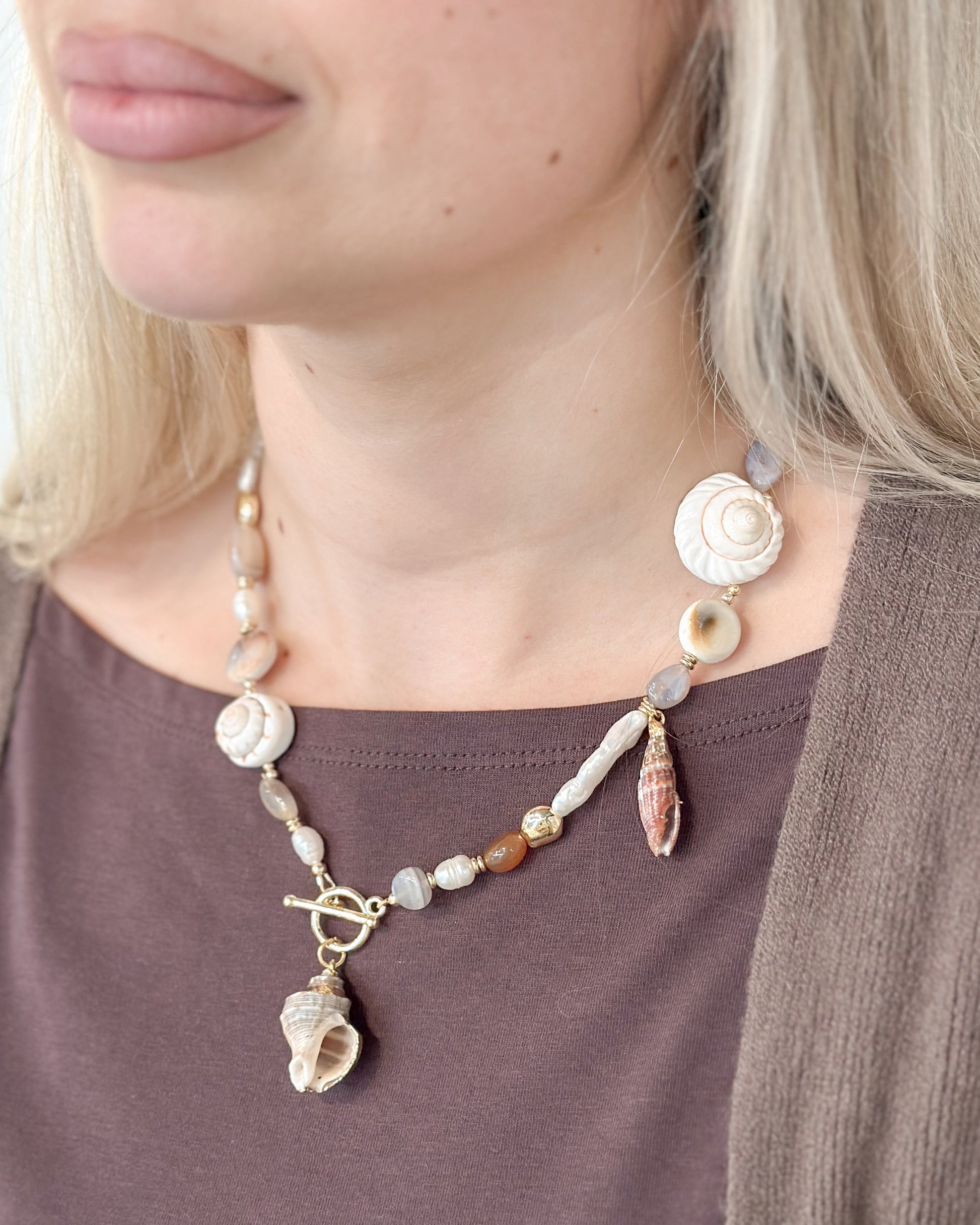 A close-up of a woman wearing a bohemian-style seashell necklace with a toggle clasp over a brown top and cardigan.