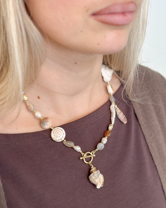 A close-up of a woman wearing a bohemian-style shell and bead necklace with a gold clasp and a conch shell pendant, paired with a brown top.