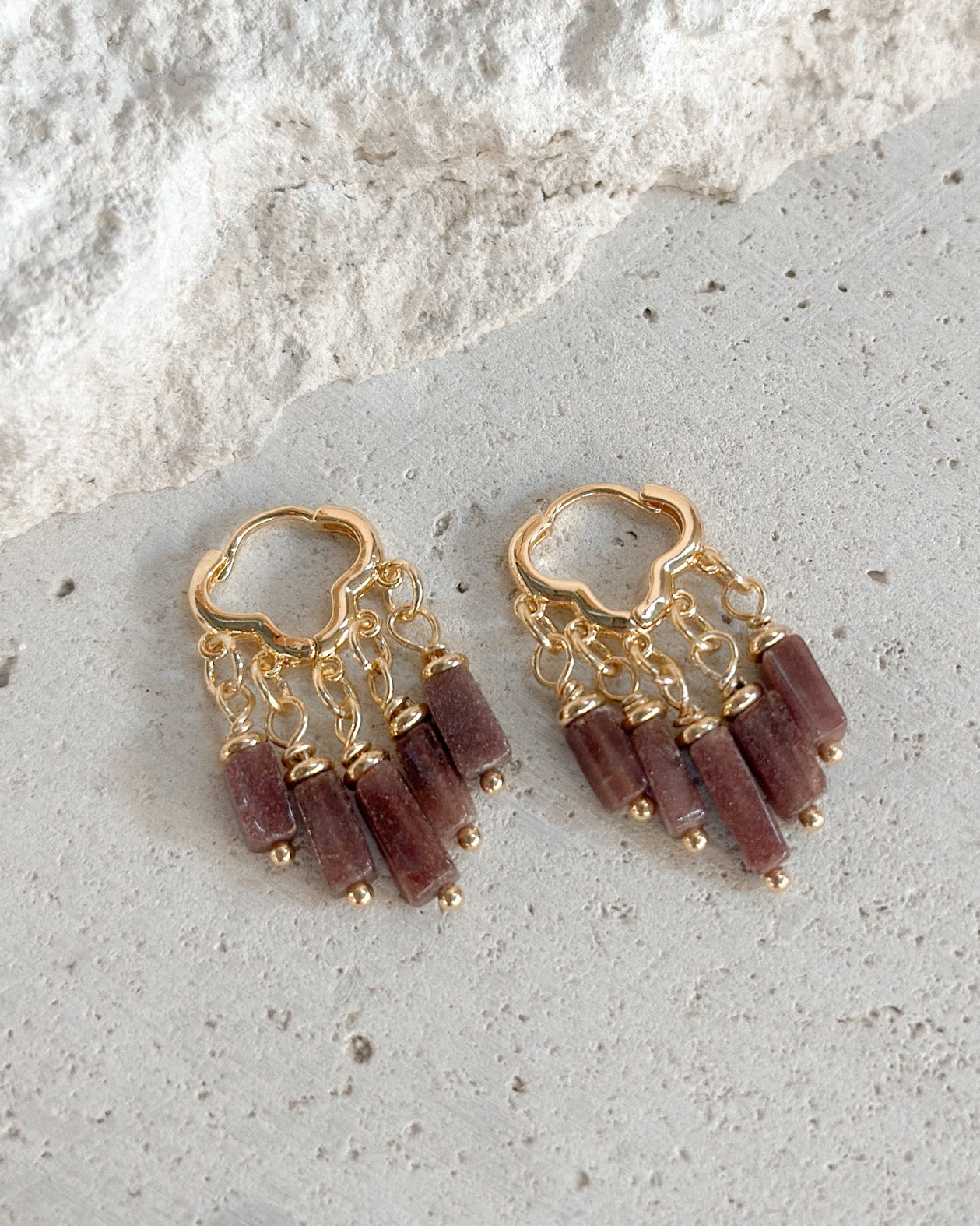 A pair of gold-toned dangle earrings with multiple small cylindrical beads in a muted burgundy or mauve tone, displayed on a light textured stone surface.