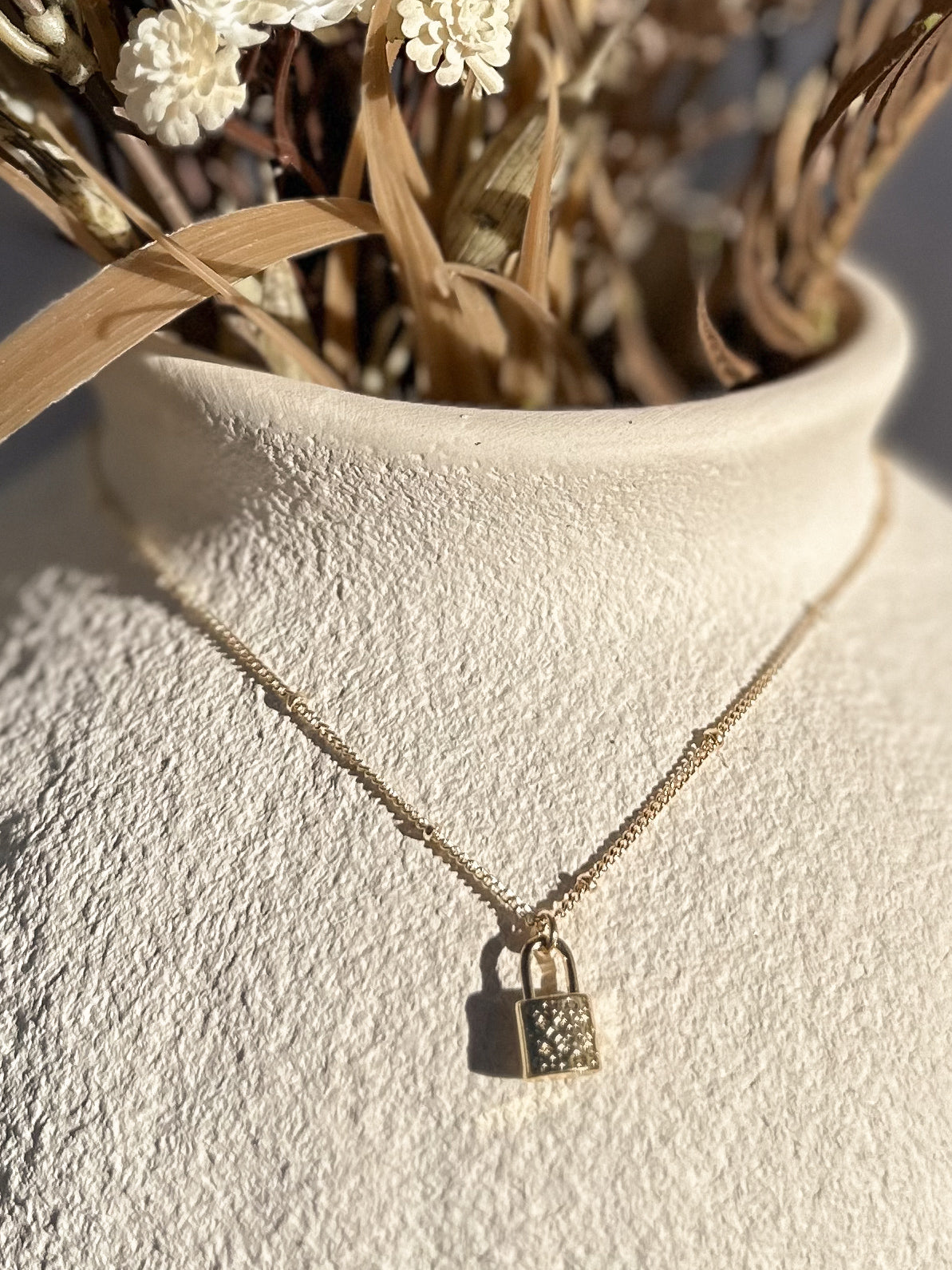 Gold padlock necklace on a textured vase with dried florals
