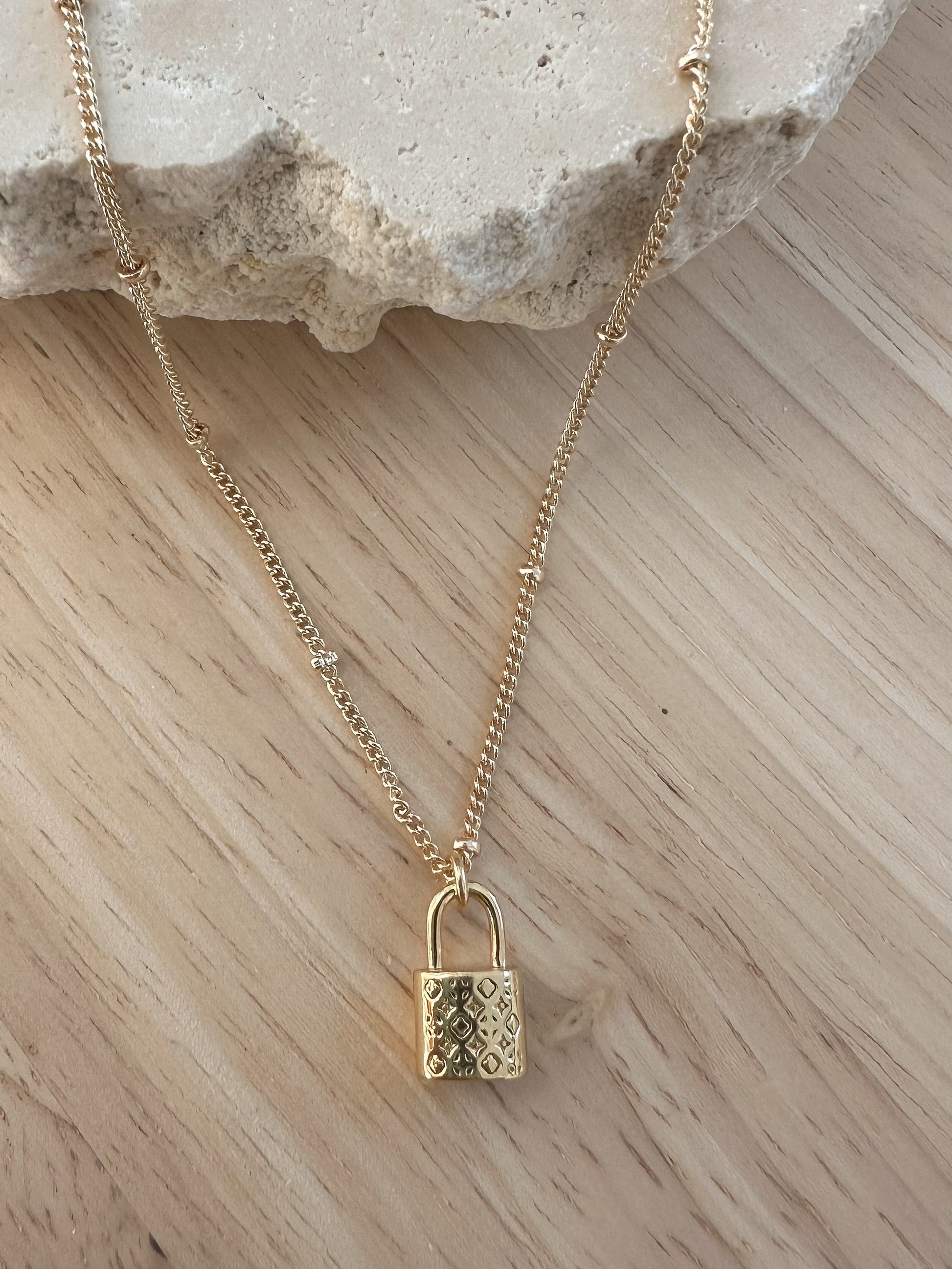 Gold padlock necklace with ornate pattern detailing draped over a travertine stone on a timber background