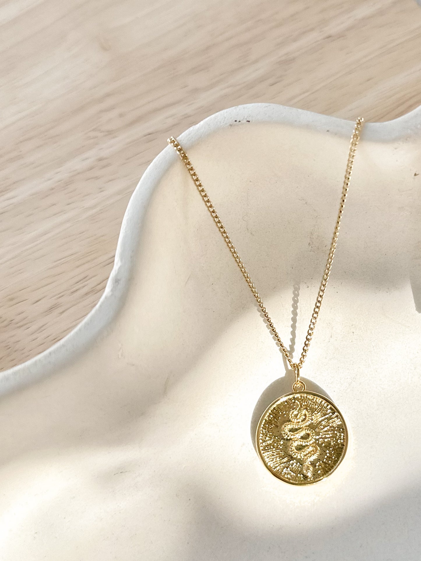 Gold sunburst serpent pendant necklace displayed on textured ceramic vase.