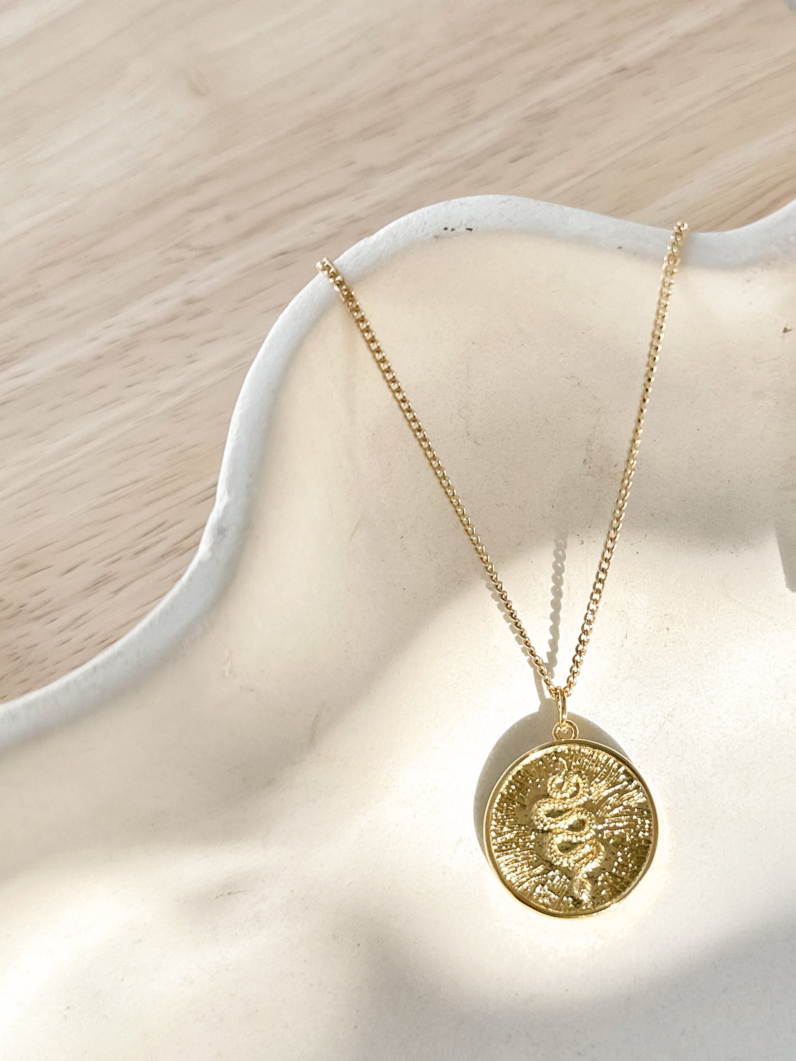 Gold sunburst serpent pendant necklace displayed on textured ceramic vase.