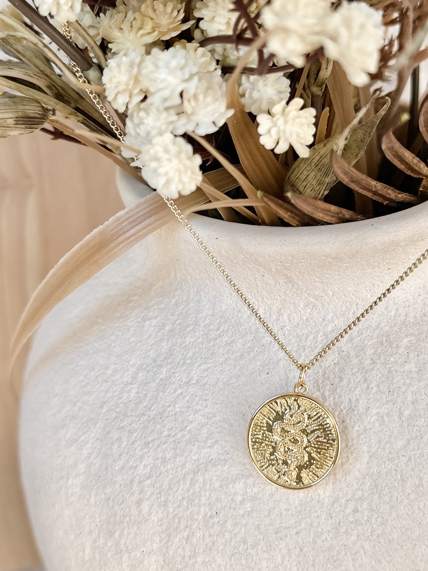 Gold sunburst serpent pendant necklace displayed on textured ceramic vase.