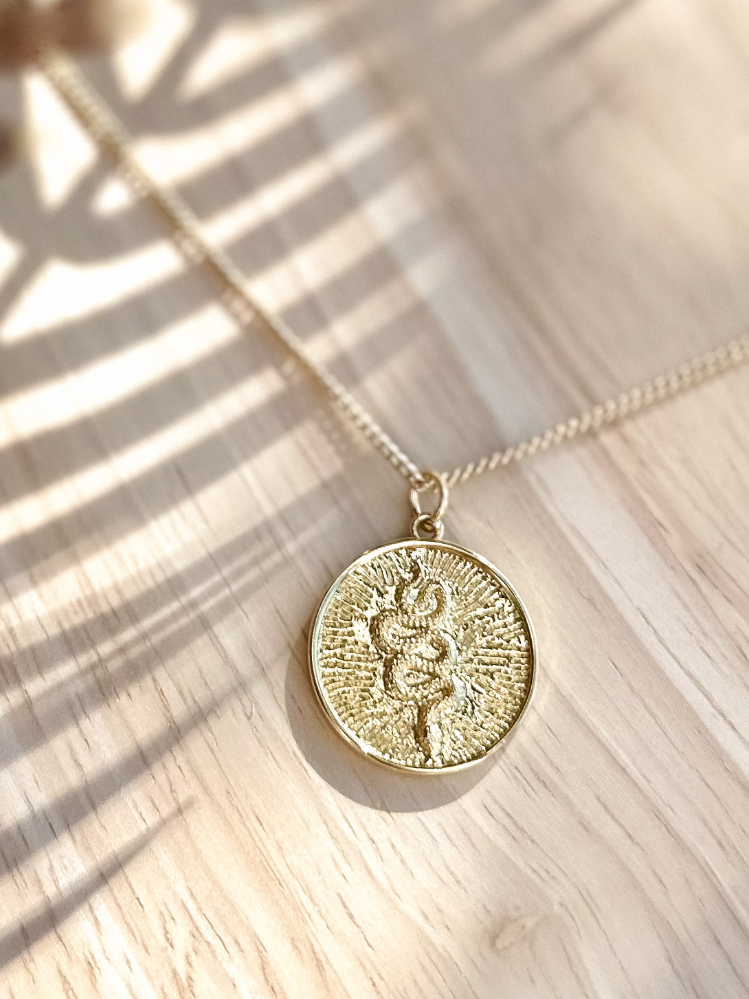 Gold sunburst serpent pendant necklace displayed on timber surface with soft palm leaf shadows