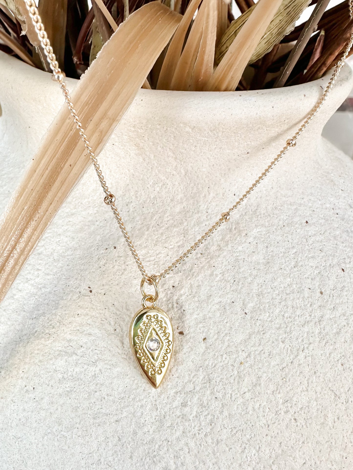 Gold necklace with a decorative pendant on a textured surface with dried plants in the background