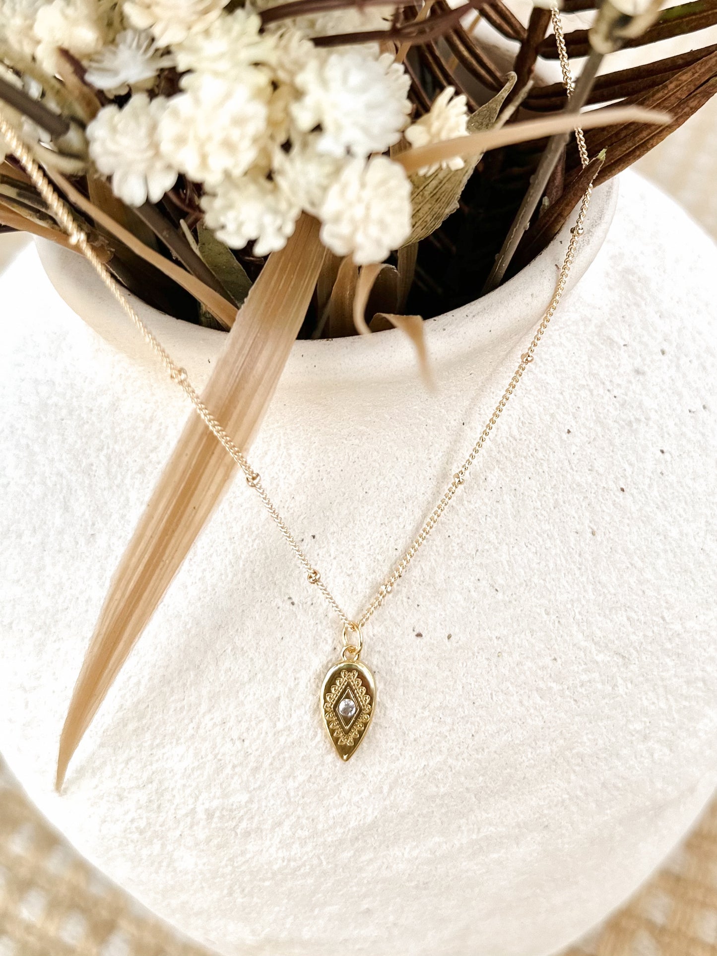 Gold necklace with a pendant on a white textured surface with dried flowers.