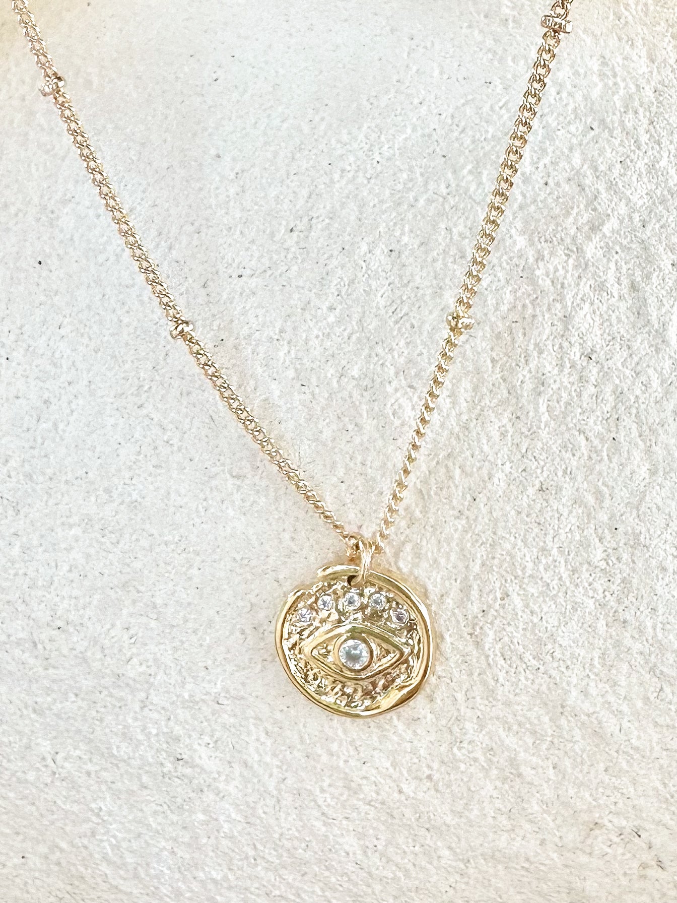Gold round evil-eye pendant necklace with cubic zirconia displayed on ceramic surface.