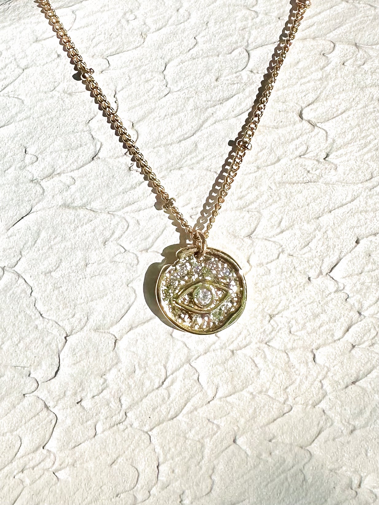 Gold round evil-eye pendant necklace with cubic zirconia displayed on ceramic surface.