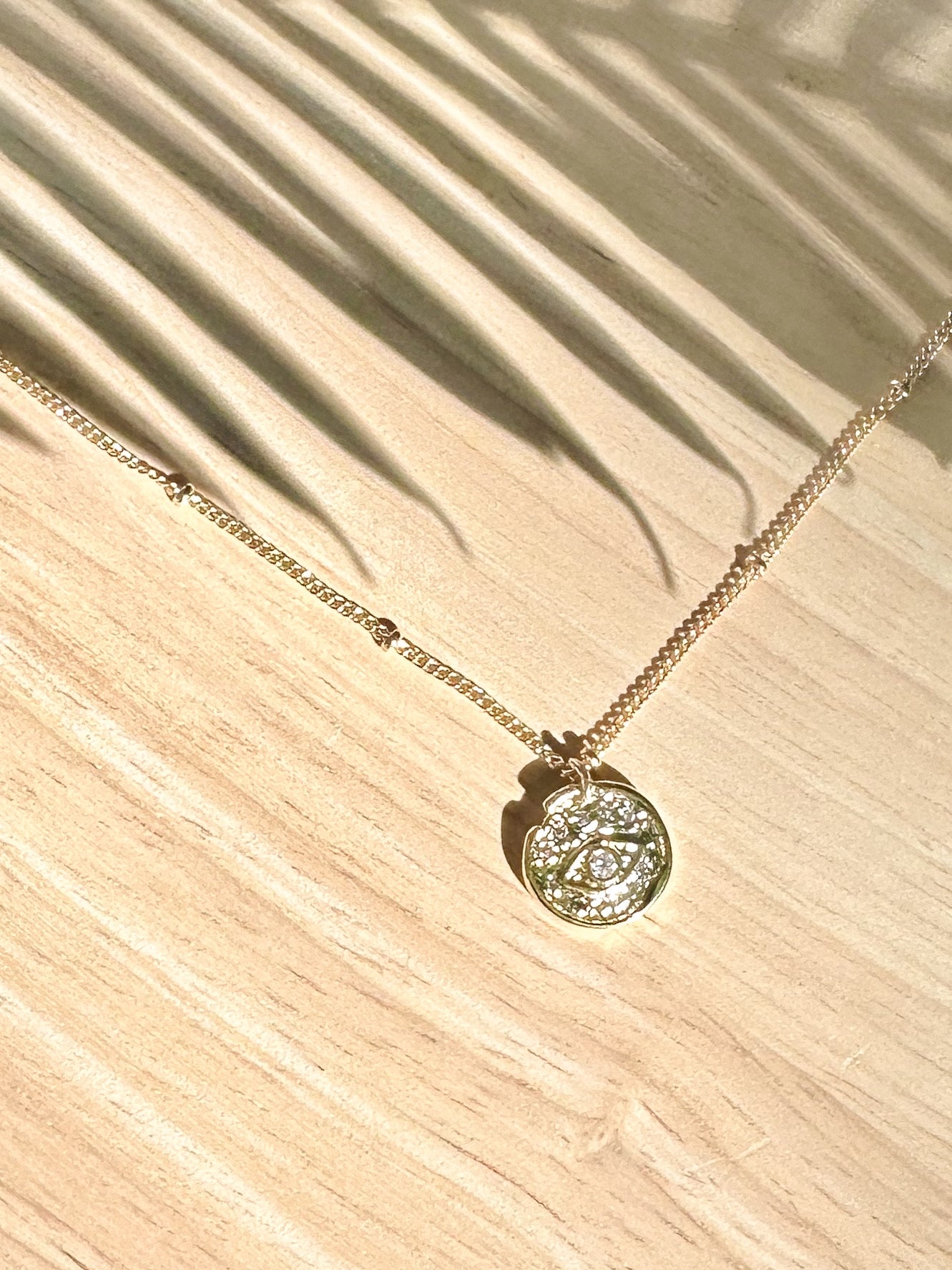Gold round evil-eye pendant necklace with cubic zirconia displayed on timber surface with palm leaf shadows