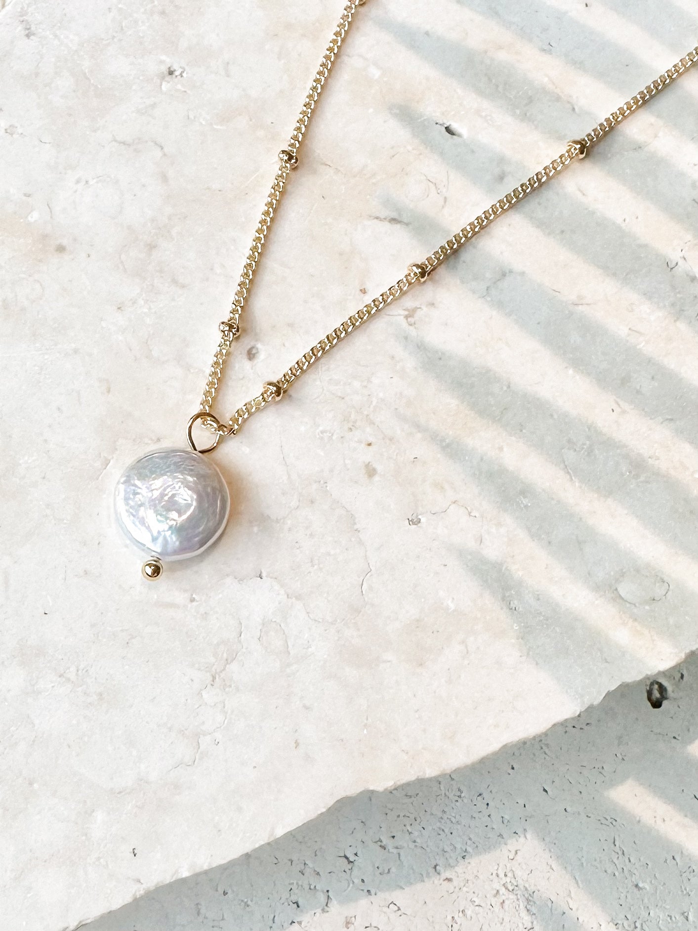 Gold coin pearl necklace displayed against soft stone background with palm shadows