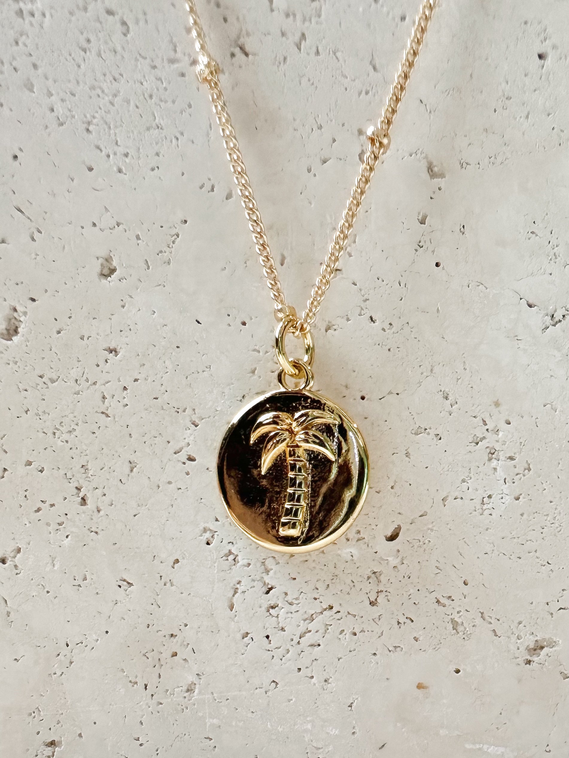 Gold necklace with a palm tree pendant on a travertine stone background
