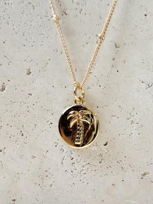 Gold necklace with a palm tree pendant on a travertine stone background