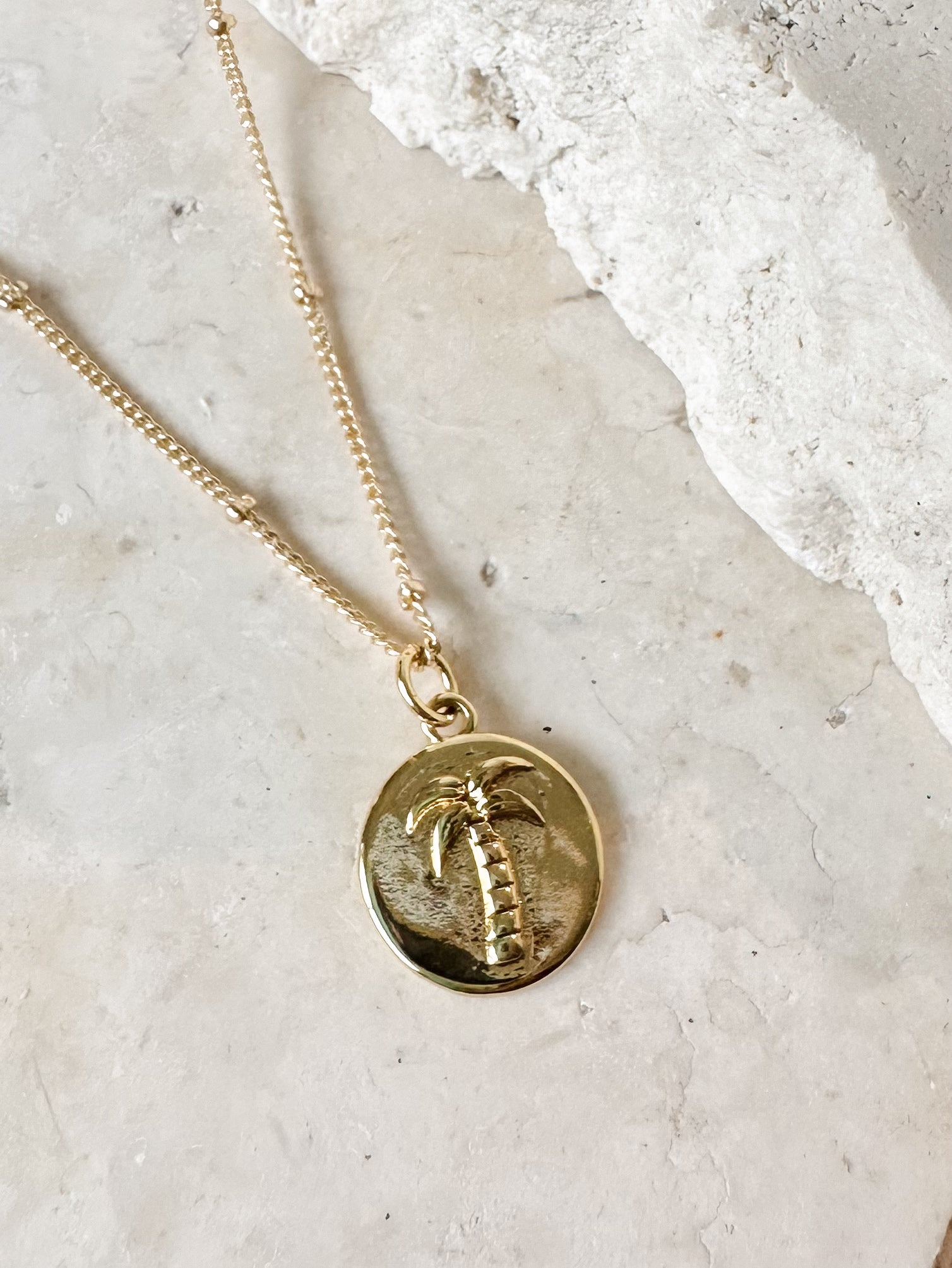 Gold necklace with a palm tree pendant on a light stone surface