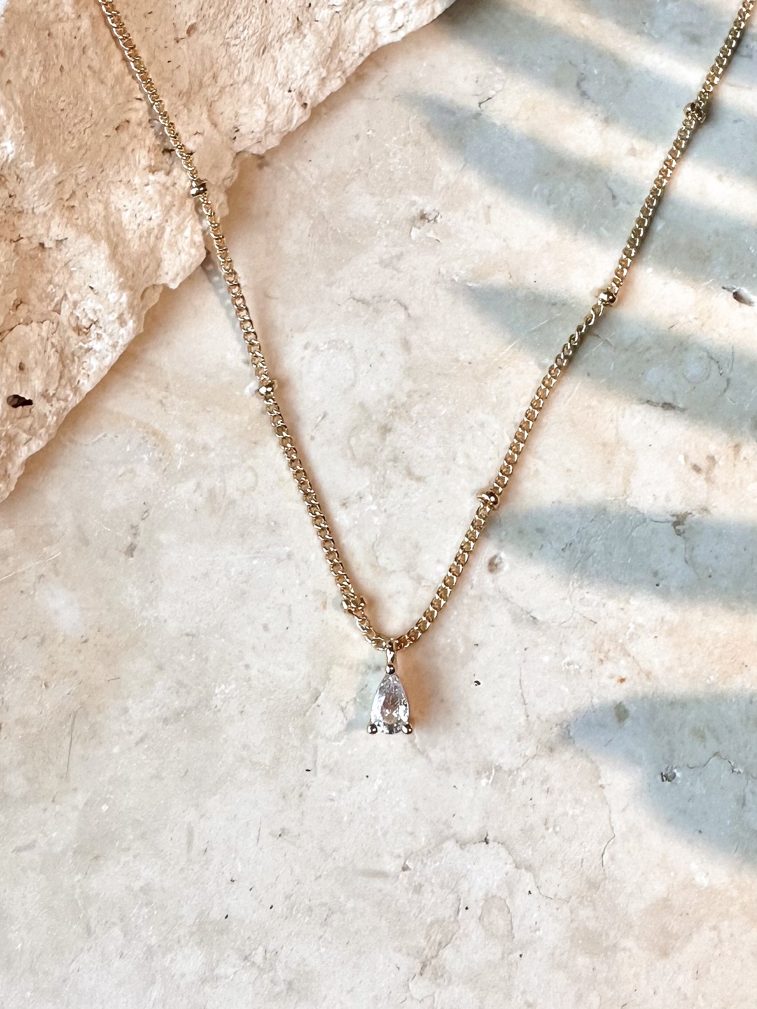 Gold necklace with a clear cz pear shaped pendant on a travertine stone surface