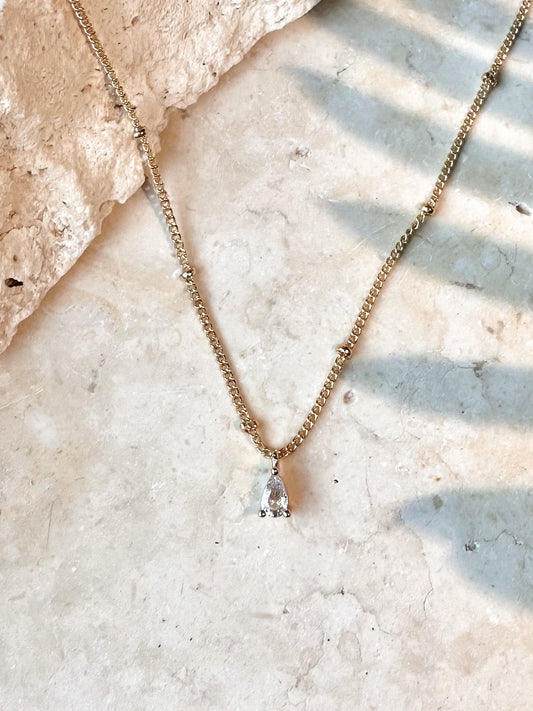 Gold necklace with a clear cz pear shaped pendant on a travertine stone surface