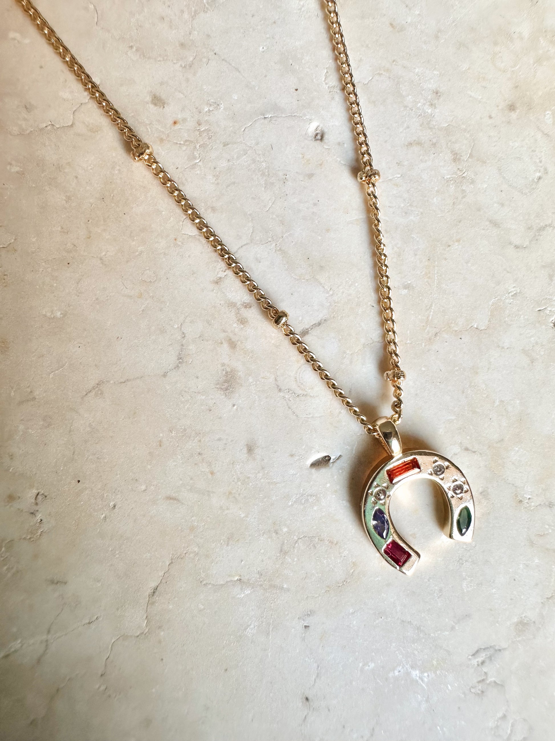 Gold necklace with a colourful cz horseshoe pendant on a light marble background