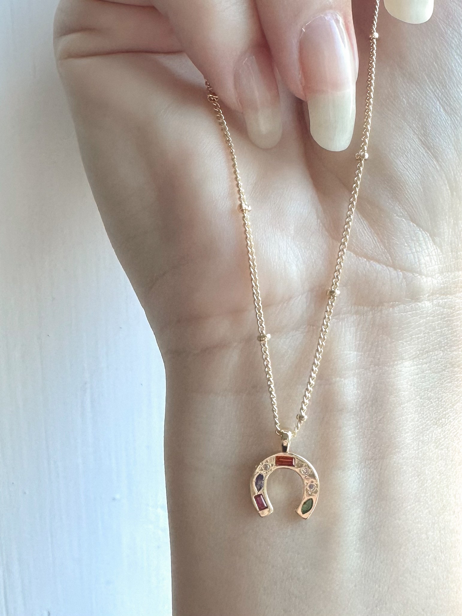 Hand wearing a gold necklace with a horseshoe pendant on a light background