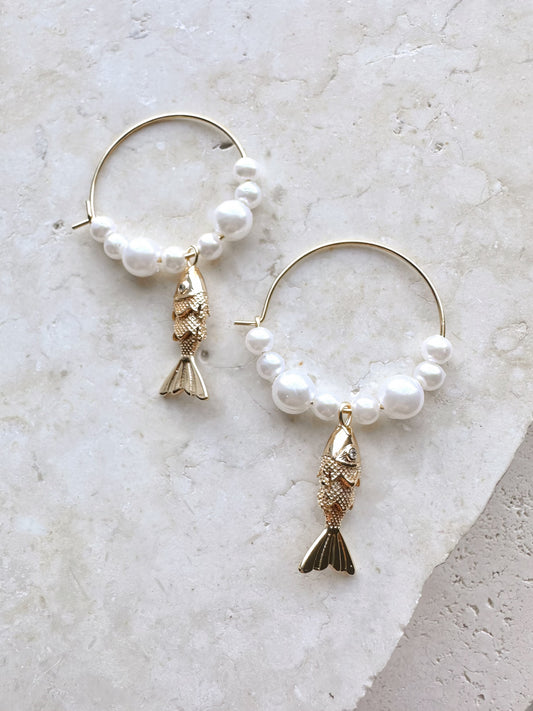 Pearl hoop earrings with gold fish charms on a travertine stone surface
