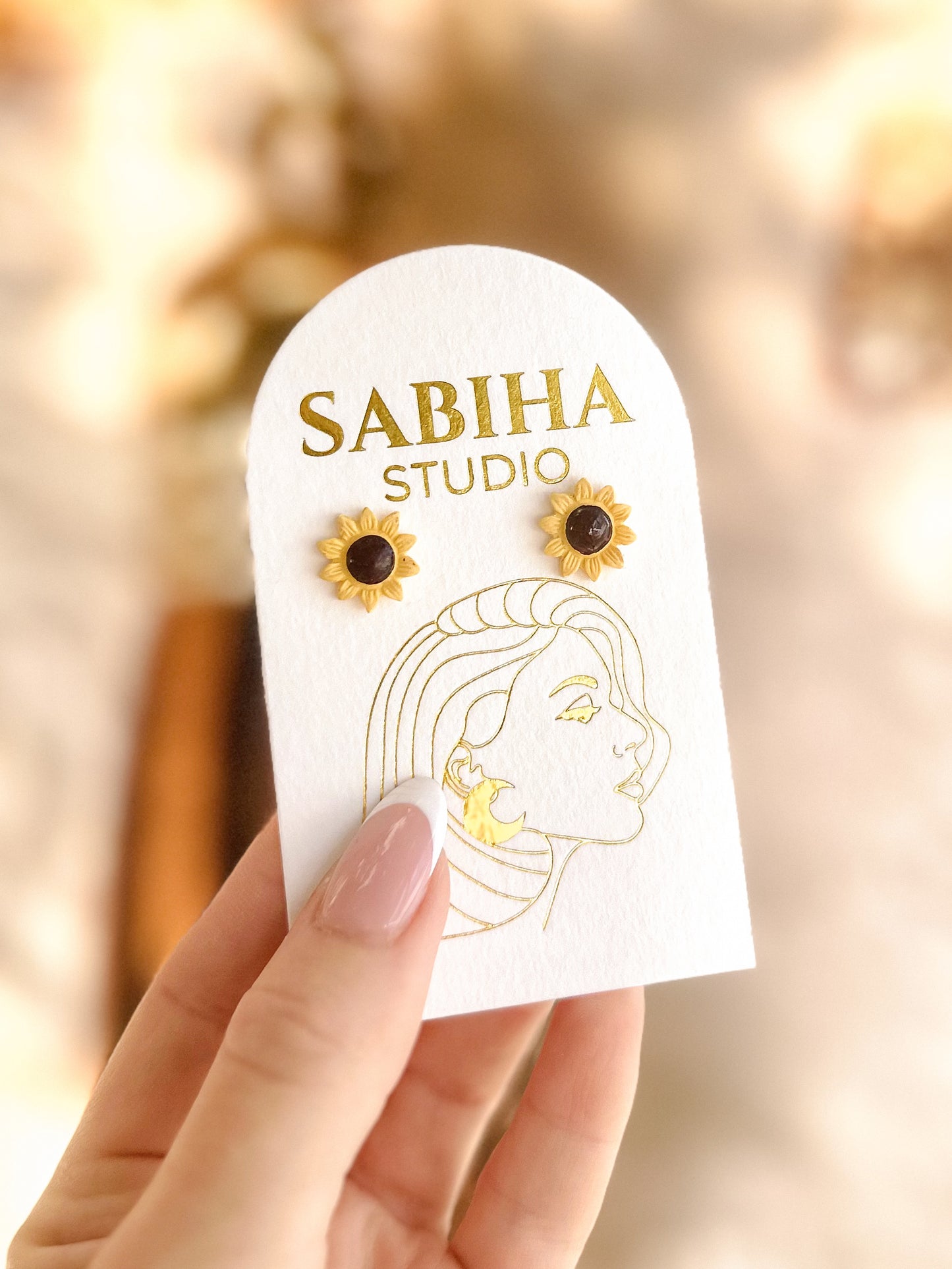 Sabiha Studio yellow sunflower earrings with a black centre on a white card held by a hand with a blurred background