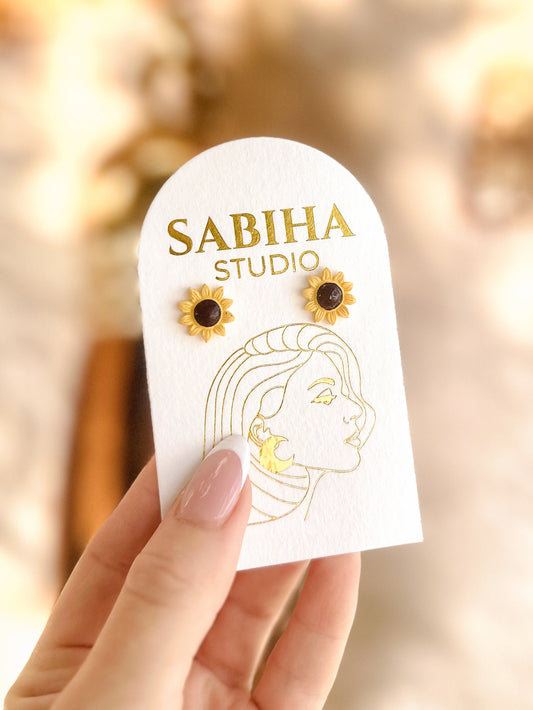 Sabiha Studio yellow sunflower earrings with a black centre on a white card held by a hand with a blurred background
