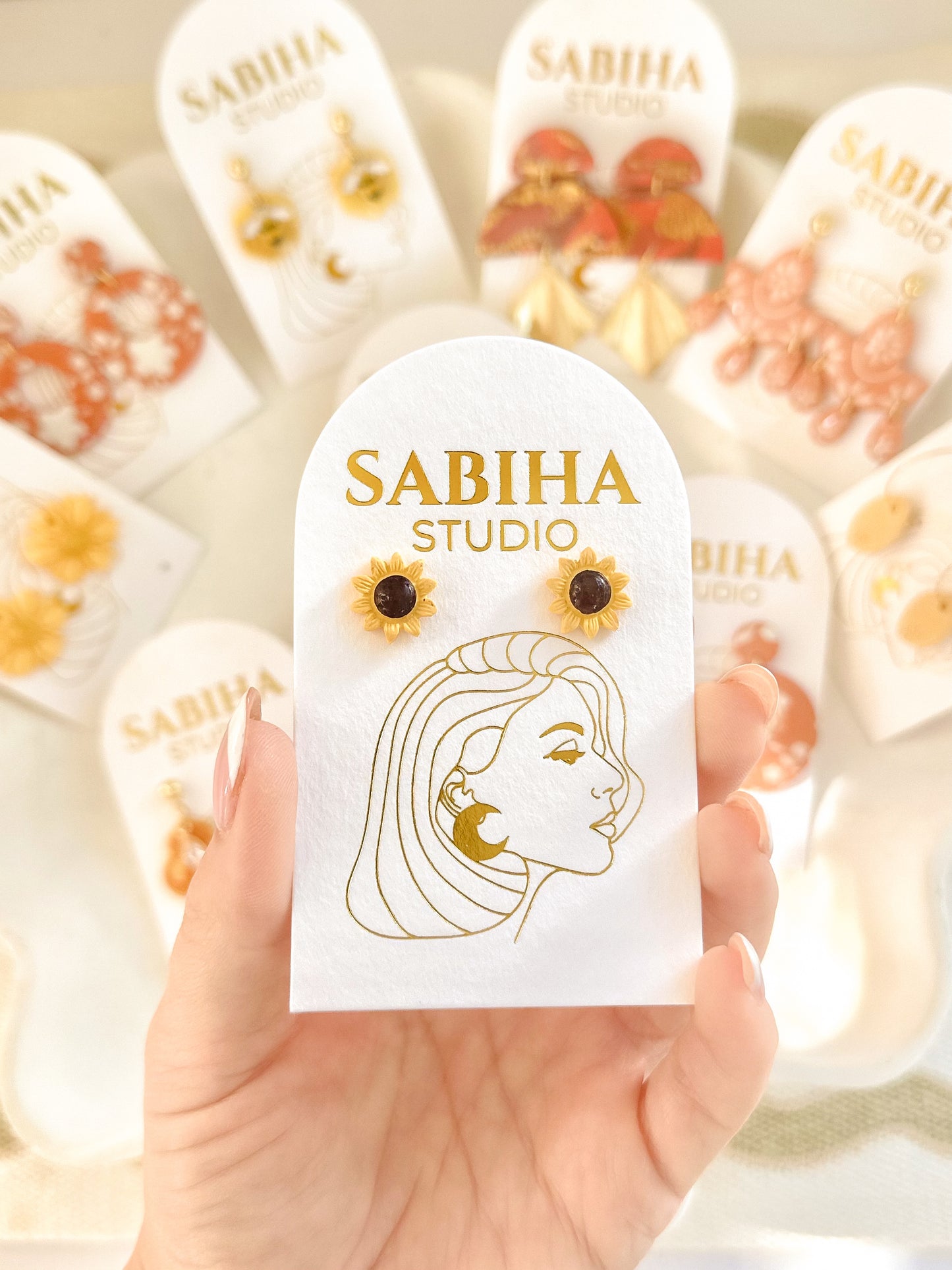 Sabiha Studio yellow and black sunflower stud earrings displayed on a card with a hand holding it.