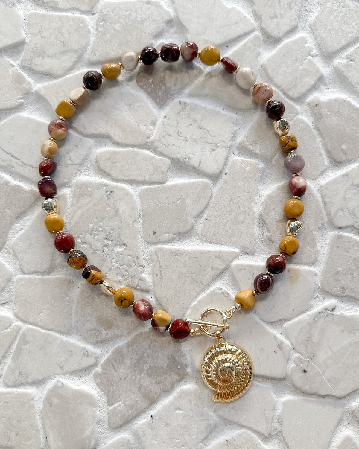 A beaded necklace with a gold-toned spiral shell charm, displayed on a light gray stone tile surface. The beads feature a mix of earthy colors including yellow, brown, red, and pale pink.