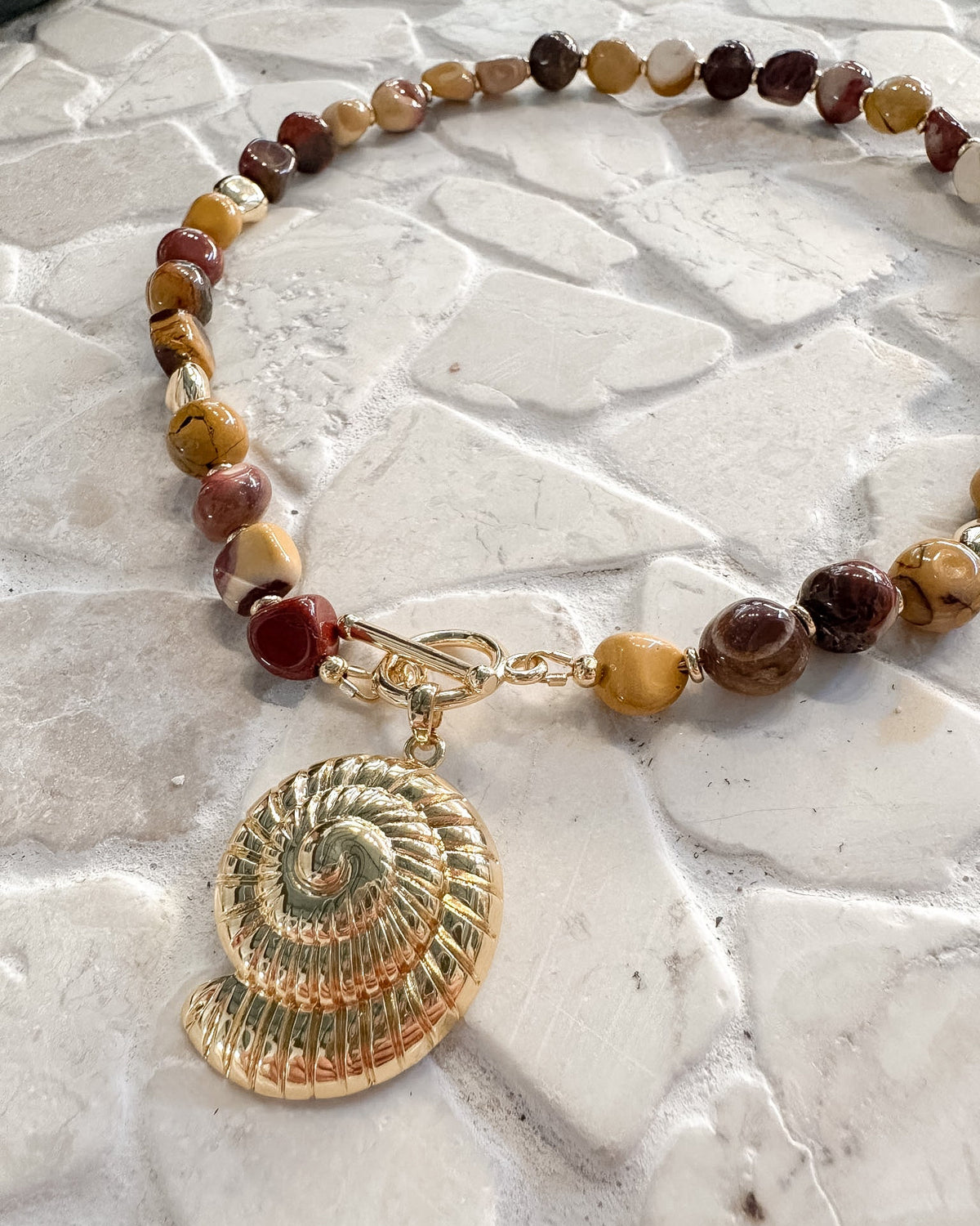 A beaded necklace featuring natural stone beads in earthy brown, red, and yellow tones, with a gold-tone shell-shaped charm and clasp, displayed on a light stone-textured surface.