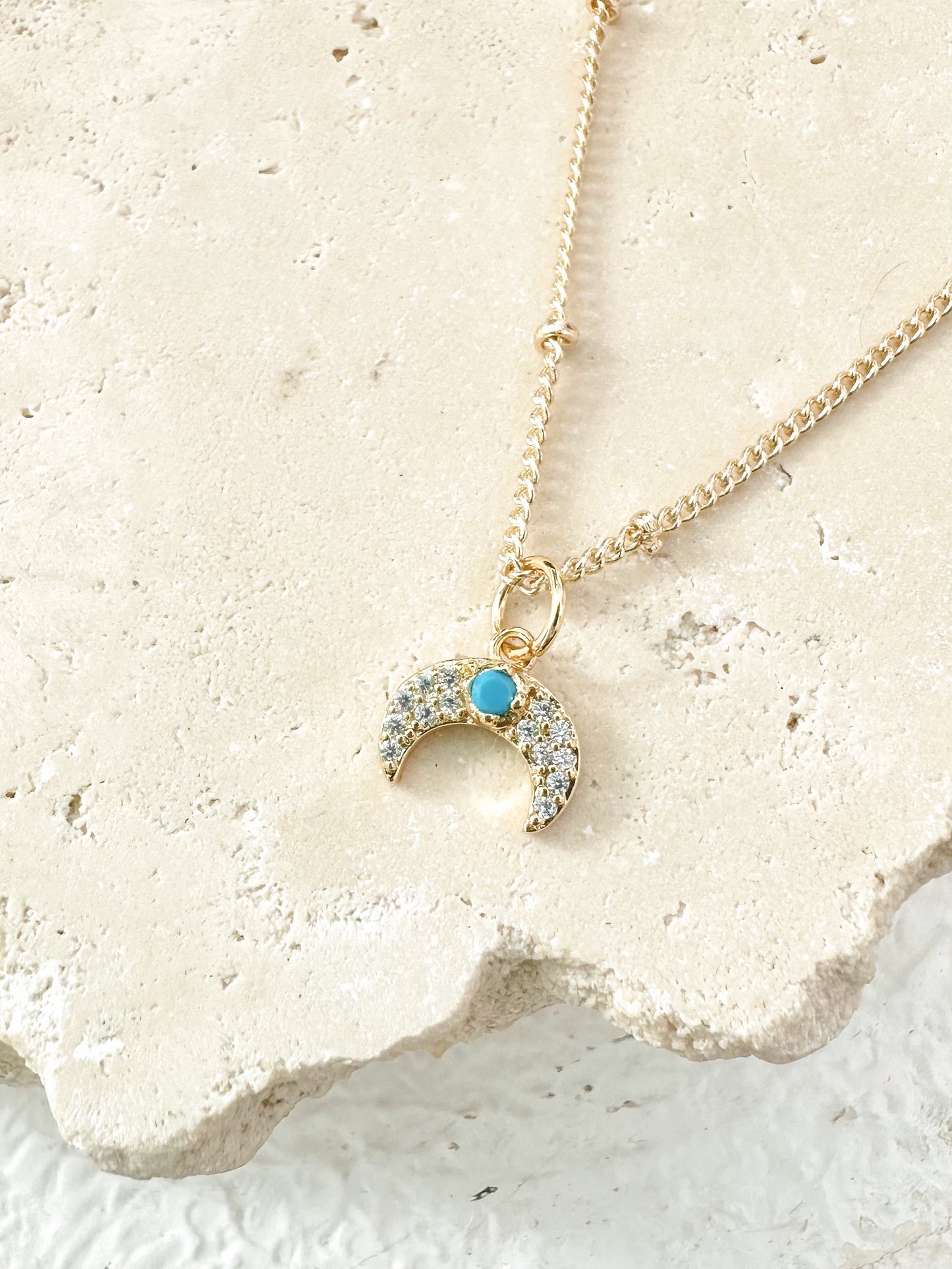 Gold crescent moon necklace with turquoise centre and cubic zirconia displayed on travertine.